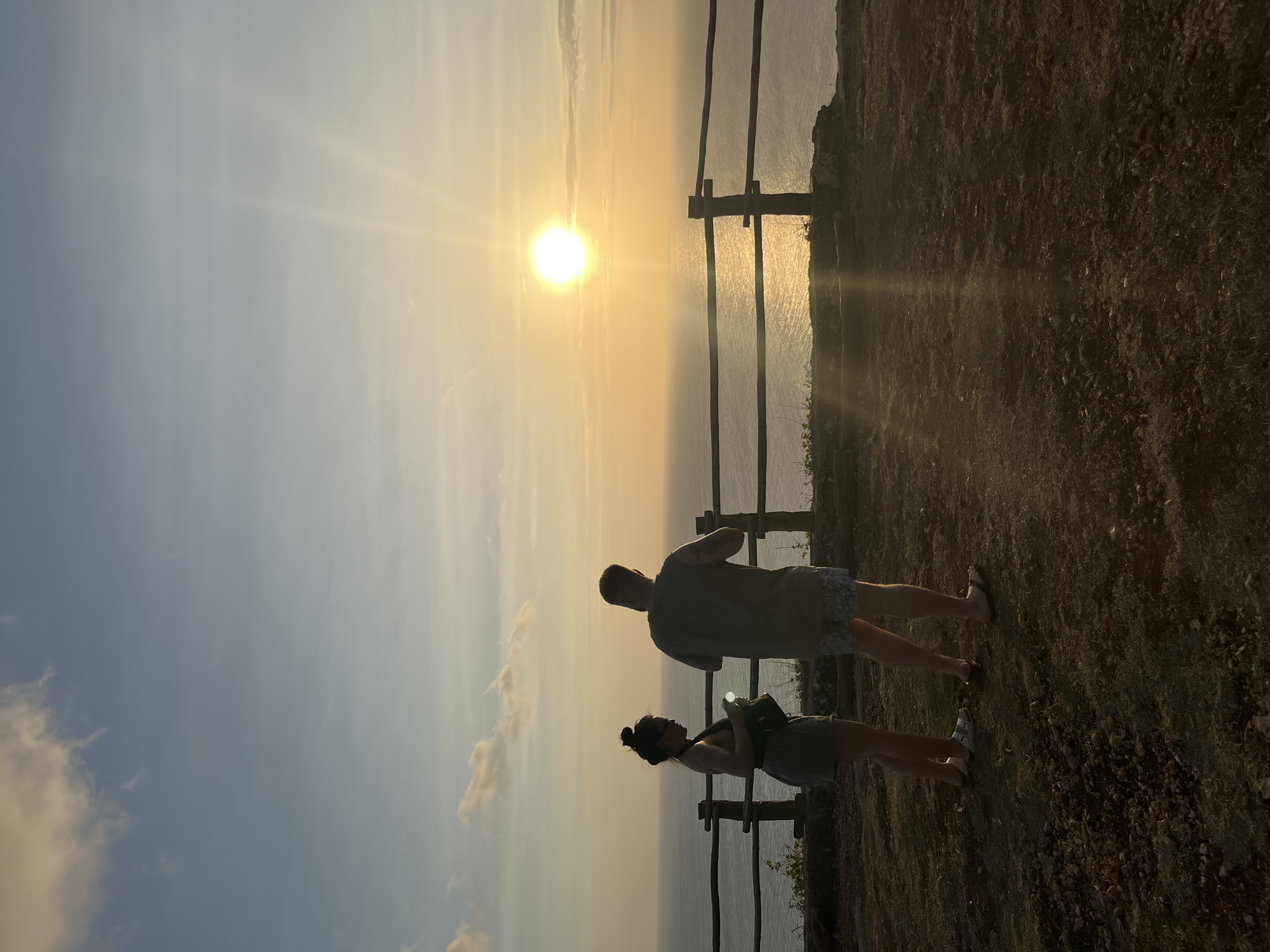 Two individuals, silhouetted against a vibrant sunset over the ocean, stand on a clifftop overlooking the water. A wooden railing separates the clifftop from the steep drop to the sea. The man, on the right, wears patterned shorts and a t-shirt, with his back to the camera. The woman, on the left, faces slightly away, wearing a dark tank top and shorts, holding a small object in her hands. The sun, a bright orb, is low on the horizon, casting a golden glow on the water and creating visible light rays. The sky is a gradient of blue and orange, with scattered clouds. The foreground consists of dry, uneven ground with sparse vegetation. This scene is consistent with a viewpoint at Uluwatu Temple in Kuta Selatan, Bali, Indonesia, a popular location for watching the sunset.
