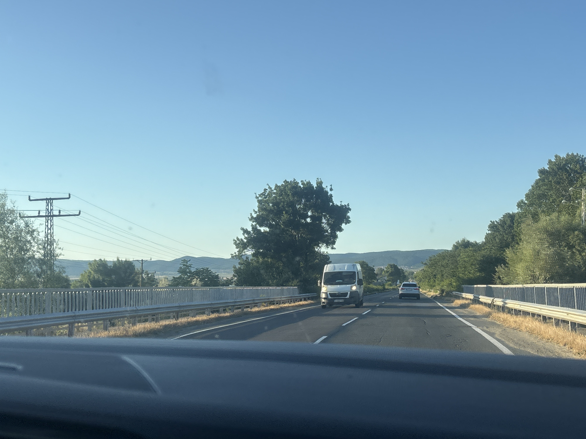 The image depicts a highway scene on a clear, sunny day. The viewpoint is from inside a vehicle, looking forward. The road is asphalt with white lane markings. On the left side of the road, there is a metal guardrail separating the highway from an embankment with dry grass. A large, dark green tree stands prominently on the left side of the road, behind the guardrail. Further to the left, a tall electricity pylon with wires is visible. In the distance, rolling hills covered in vegetation can be seen.
On the highway, two vehicles are present. A white van is in the right lane, traveling away from the camera. Its front end and headlights are clearly visible. Further ahead in the right lane, a silver car is also visible. The right side of the highway also has a metal guardrail with an embankment of dry grass behind it. The sky is a clear, light blue.
Based on the provided information, this scene is located on a highway near Nesebar, Bulgaria.