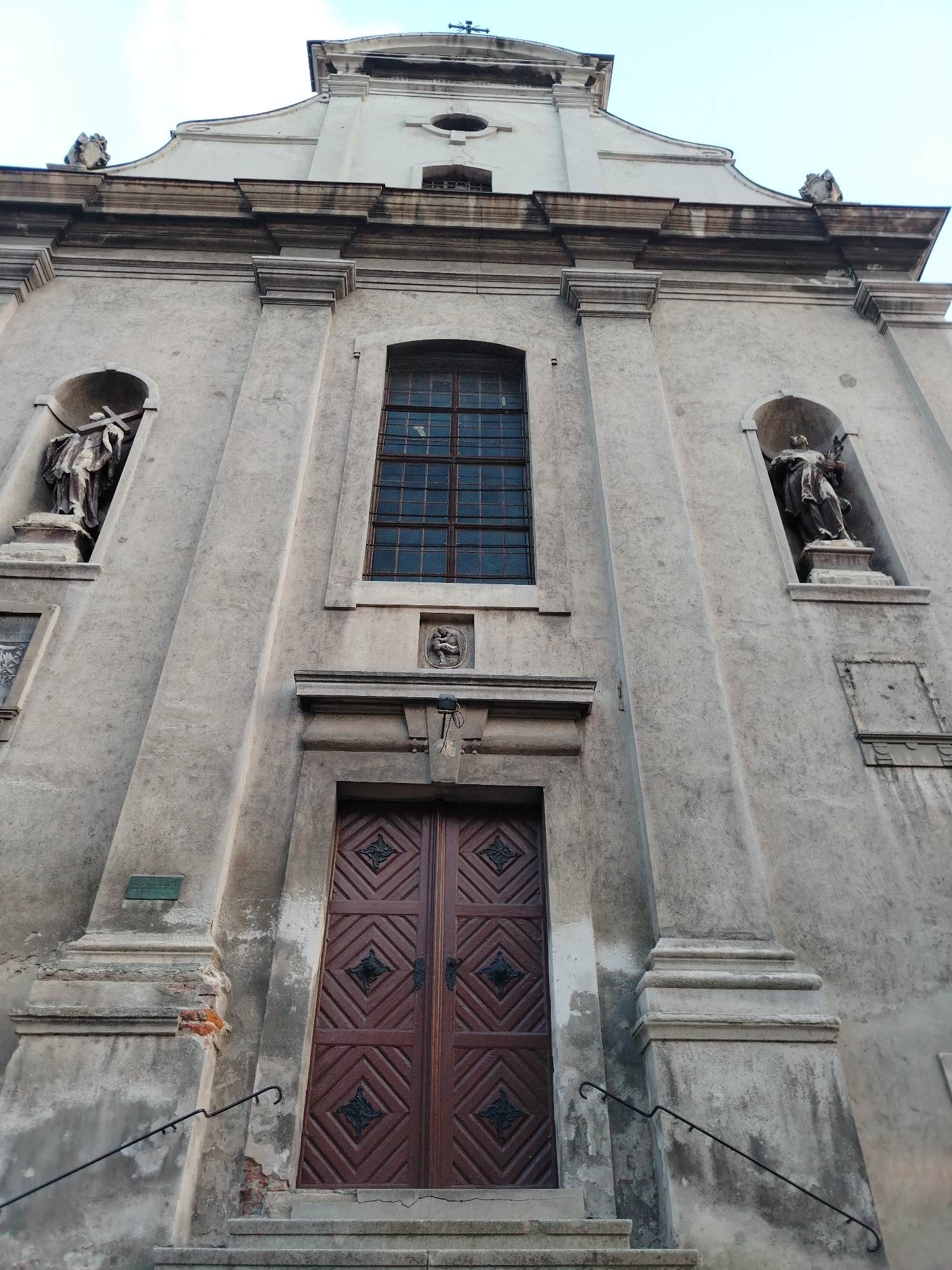 This is a low-angle, eye-level, full-shot photograph of the facade of a stone church. The church is an old, ornate building with statues and intricate carvings. The sky is a pale blue. The church has a large, dark wooden door with diamond patterns and ornate metal hinges. To the left and right of the door are two arched niches, each containing a statue of a robed figure holding a cross. Above the door is a small, circular carving. A tall, narrow window with a dark metal grate is positioned above the door and carving. The facade is made of rough, weathered stone and shows signs of age and deterioration. There are two staircases leading up to the door, with simple black metal railings. The overall impression is one of historical grandeur and religious significance. Based on the architectural style and the mention of Petrovaradin, Serbia, this is likely a religious building within the Petrovaradin Fortress.