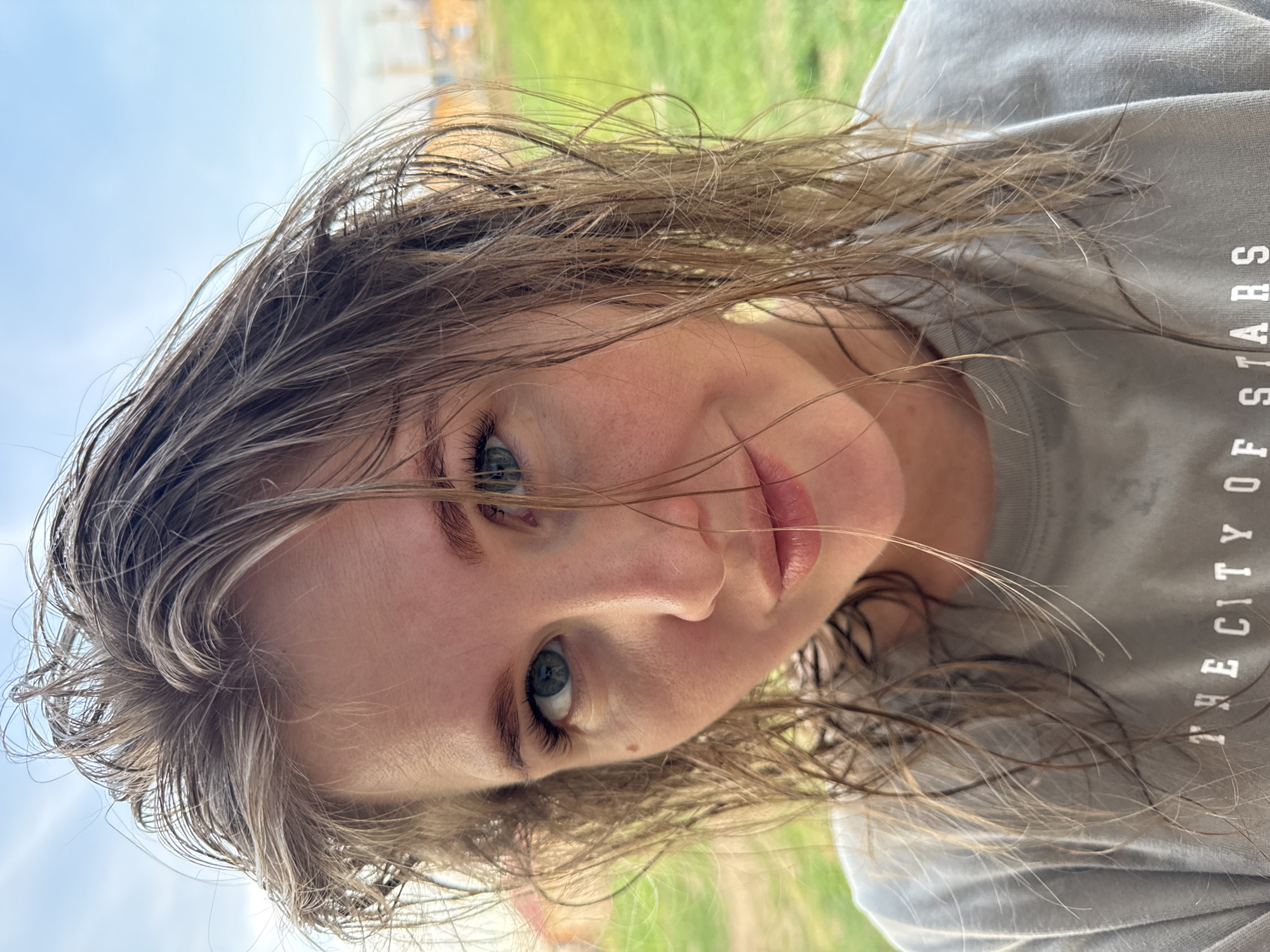 A close-up shot shows a woman with wet, light brown hair. She has fair skin, blue eyes, and light pink lips. She is wearing a light gray T-shirt with the text "THE CITY OF STARS" partially visible. The background appears to be outdoors with green grass and blue sky partially visible. The location is Ферма, Russia.