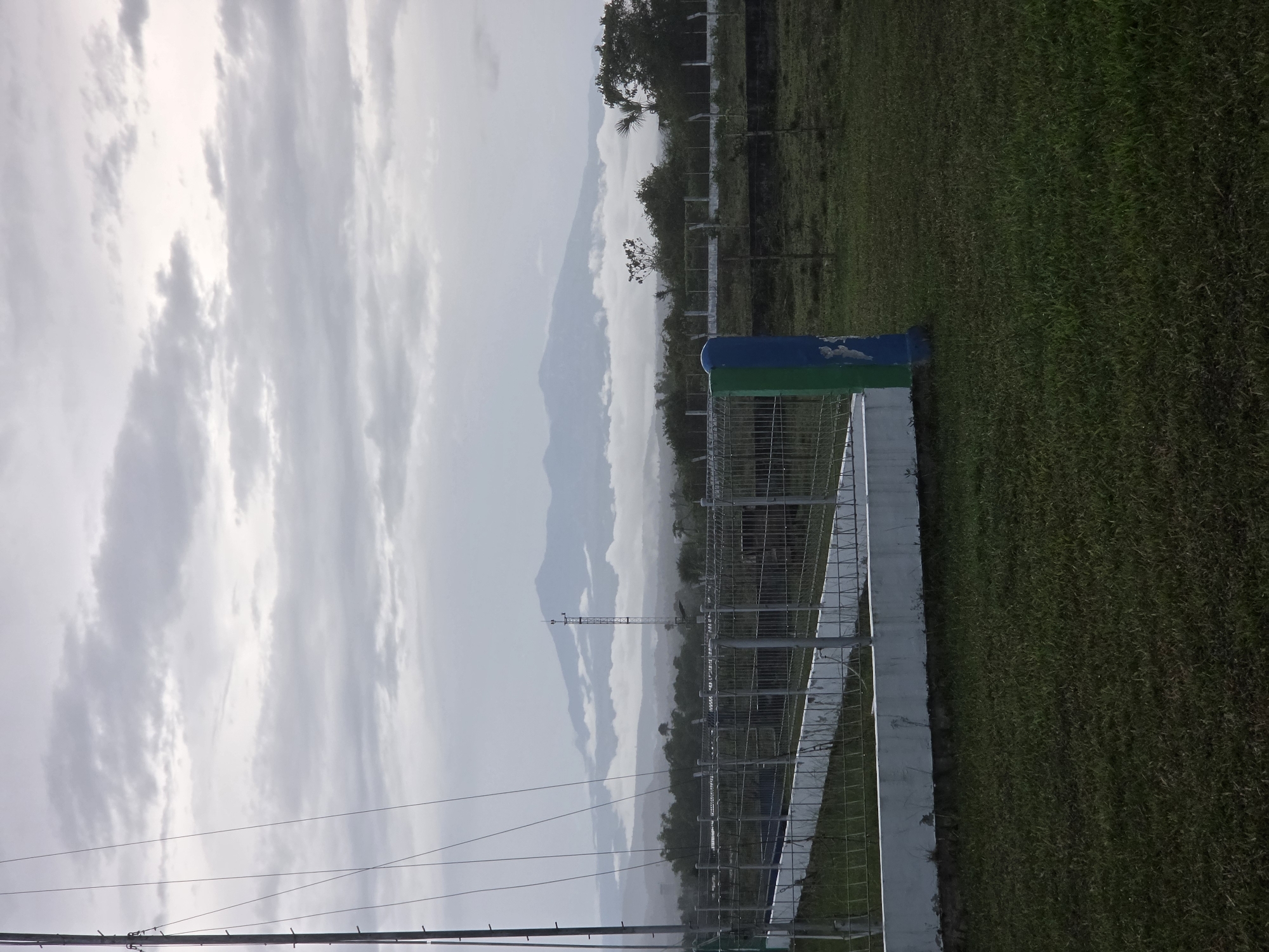 The image shows a landscape in Pintomakmur, Indonesia. A wire fence runs along the right side of the frame. Beyond the fence, there is a green, grassy area. In the distance, mountains are visible through a cloudy sky. A communications tower can be seen amidst the trees. Power lines are present in the foreground against the sky.