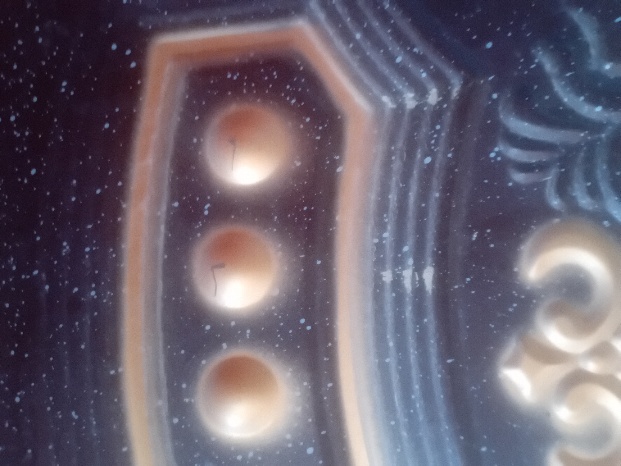 The image shows a close-up, out-of-focus view of a decorative surface, possibly a door or a piece of furniture. The surface is dark blue with scattered small white and light blue speckles, resembling stars or glitter. Prominently featured are three raised, circular elements with a golden-brown hue, each with a slightly darker, curved line visible on its surface. These circular elements are set within a hexagonal frame with a golden-brown border. Below these, further decorative elements with swirling, ridged patterns in a darker blue are visible, along with a glimpse of ornate golden scrolling at the bottom. The lighting is uneven, creating bright highlights on the raised circular parts. The image lacks any contextual information about its location within a city, nor does it depict any people, actions, or interactions.