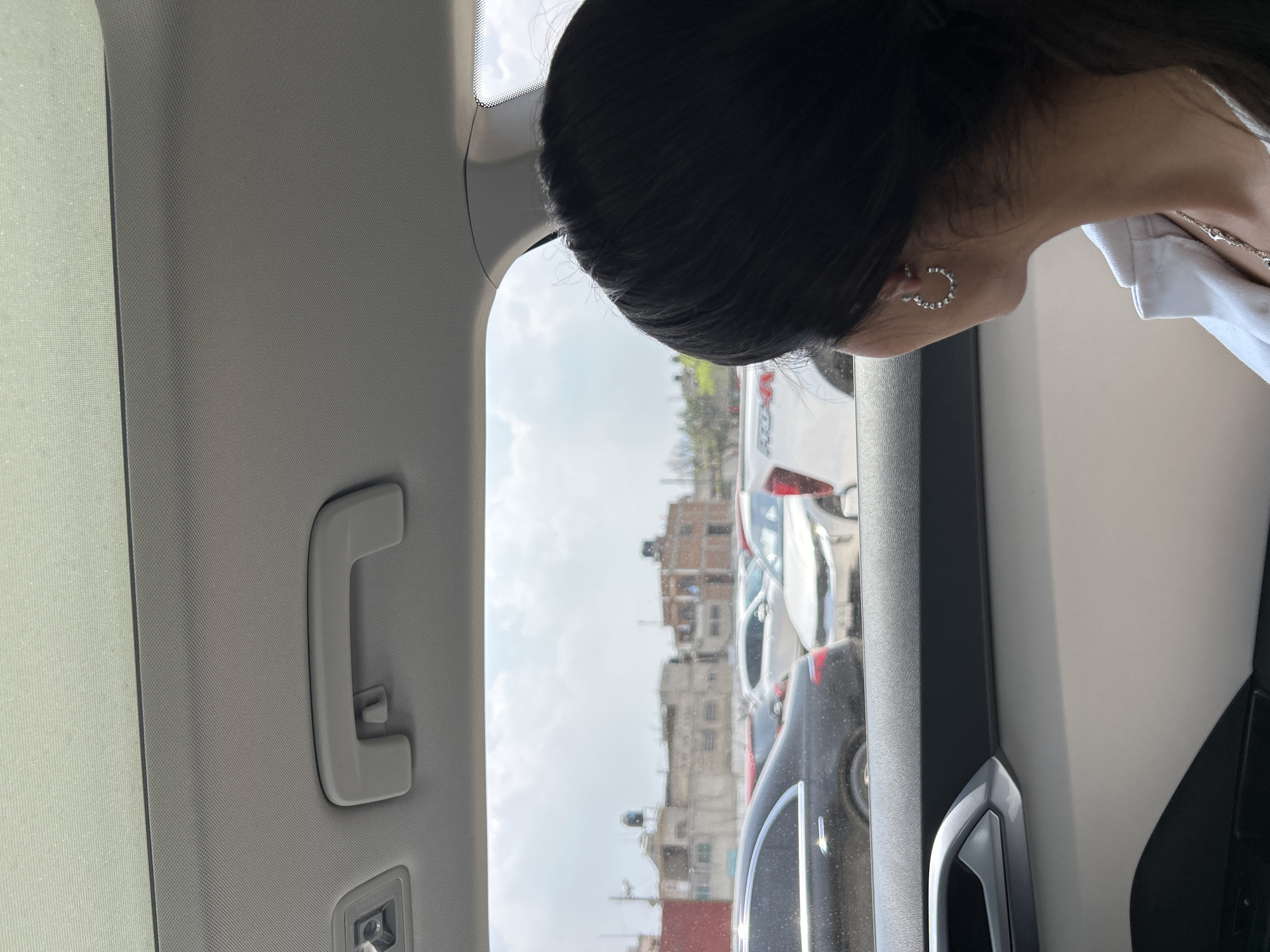 A person is seen from the neck up to the top of the head, turned toward the car window. The person has dark hair, an earring on the exposed ear, and wears a white shirt. Outside the window, a view of a parking lot containing multiple cars and buildings in the background is visible. The sky is overcast. The scene is viewed from inside the car, with the car's interior features such as the roof, side panel, and a grab handle visible. The location is in a city, but it's not discernible from the image.
