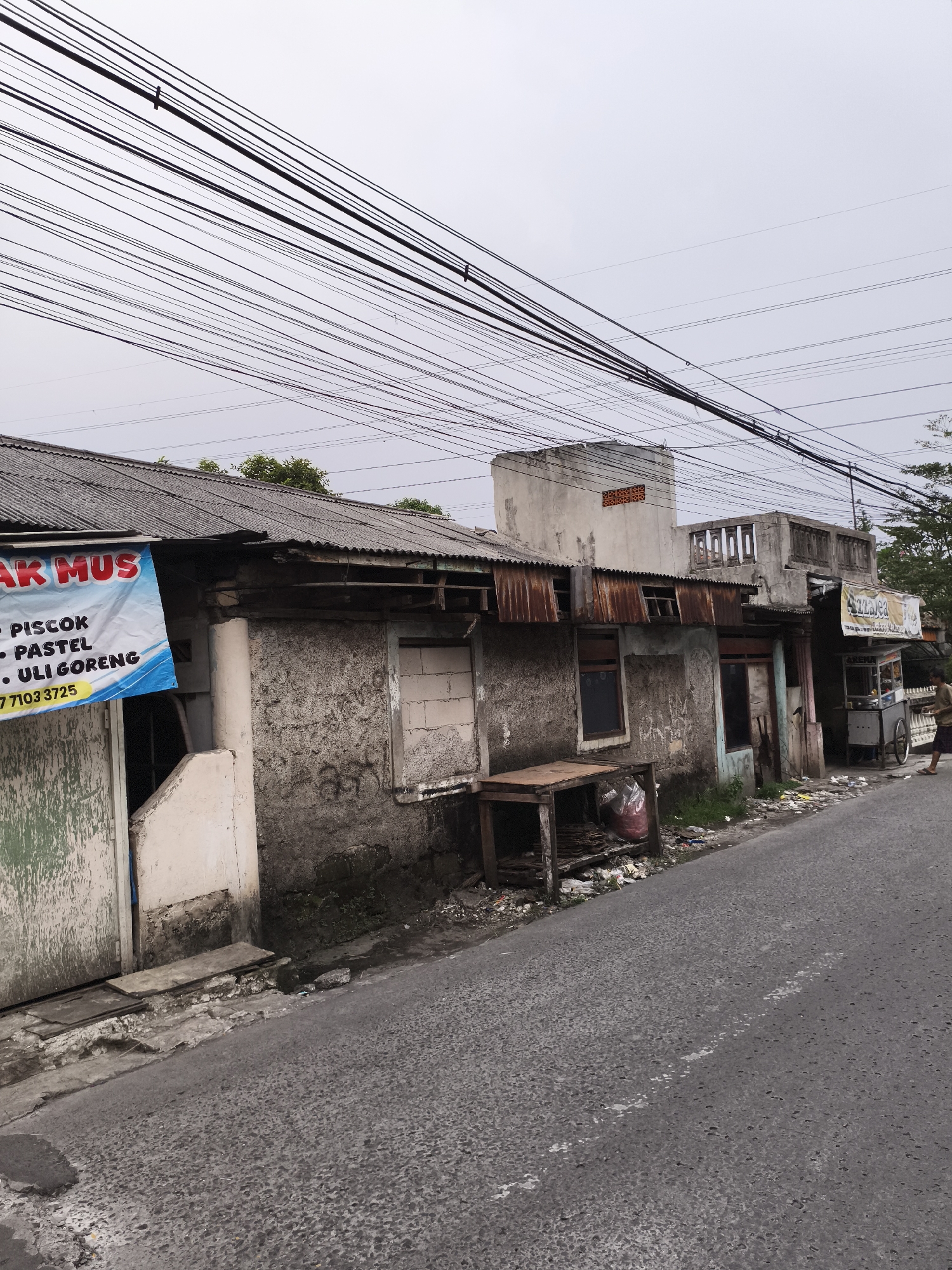 The image shows a street scene in Depok, Indonesia, characterized by modest infrastructure and overhead power lines. An aged, single-story building with a corrugated metal roof stands along the road. The building has visible signs of disrepair, with patchy concrete walls, exposed brickwork, and a partially blocked window. A blue and white sign hangs on the leftmost side of the building with the words "AK MUS" and lists of food items: "PISCOK, PASTEL, ULI GORENG" and a phone number. Adjacent to the building, a wooden table with a red plastic bag beneath it sits on the ground. To the right, a food cart labeled "AZZARICA" is visible. A person in casual attire stands near the food cart. Debris is scattered along the roadside. The street itself is paved, with visible cracks and wear. The sky is overcast.