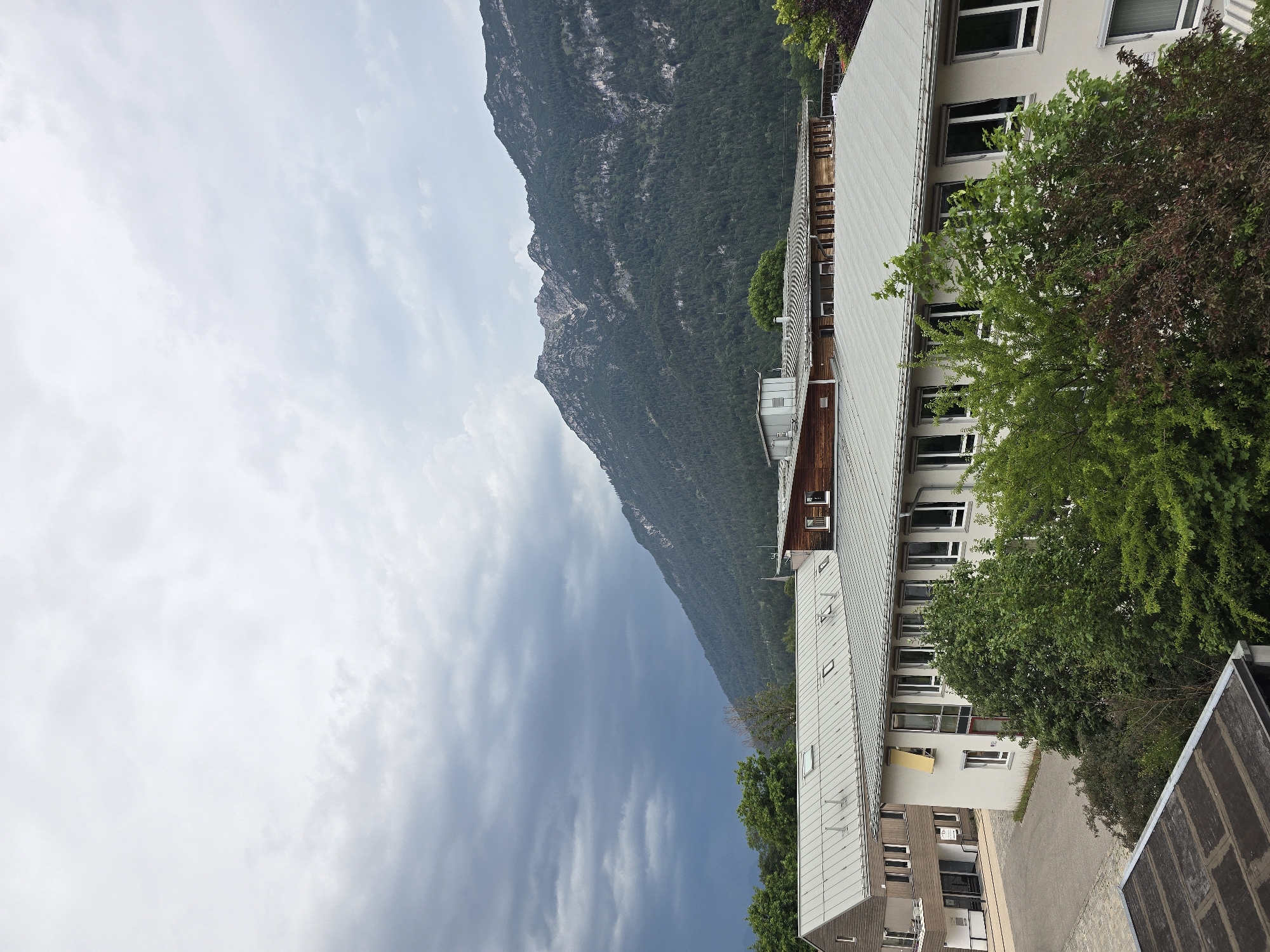 This is a photograph taken outdoors, likely in the morning or late afternoon given the soft lighting and cloudy sky. The image is oriented vertically.
The scene depicts a modern building with a light-colored, corrugated metal roof, set against a backdrop of a steep, forested mountain. The building appears to be multi-story, with numerous windows visible, some of which are open. Lush green trees, some with dark reddish-brown foliage, are interspersed around the building and in the foreground. A paved area, possibly a walkway or parking lot, is visible to the right of the building. The sky is predominantly overcast with scattered clouds, suggesting a potentially cool or temperate climate. The overall impression is one of a serene, possibly residential or commercial, setting nestled within a mountainous landscape. There are no people or discernible activities taking place in the image, focusing instead on the architectural and natural elements of the location. The specific location of Garmisch-Partenkirchen, Germany, further suggests a setting in the Bavarian Alps. No text is visible in the image.