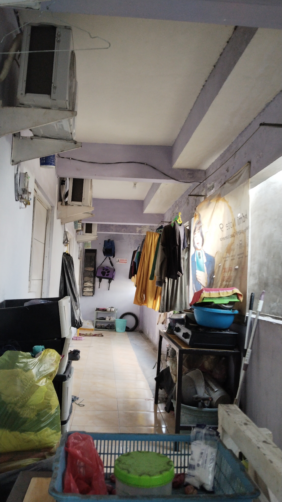 The image depicts a hallway or outdoor corridor with an overhead structure, possibly part of a residential building in Banjarsari, Indonesia. The walls are painted in a pale color with purple accents on the beams. The floor is tiled in a light beige color. On the left side, there are several air conditioning units mounted on the wall, and a white door is visible. Various items are stored and displayed, including a large yellow plastic bag, a black plastic container, and a blue plastic basket in the foreground containing a red plastic bag and a green-lidded container.
Along the right wall, clothes are hanging on a clothesline. A large banner with a person's image is also displayed. Further down the corridor, there is a shoe rack with several pairs of shoes, a bicycle tire, and a light blue bucket. A metal-framed table with a gas stove on top is positioned in the right half of the image, with various objects stored underneath. Cleaning tools, such as brooms or mops, are leaning against the wall on the far right. The lighting appears to be natural, coming from the open end of the corridor, and some artificial lights are also present. There are no people visible in the image. The scene suggests a functional, possibly utilitarian space within a residential area.