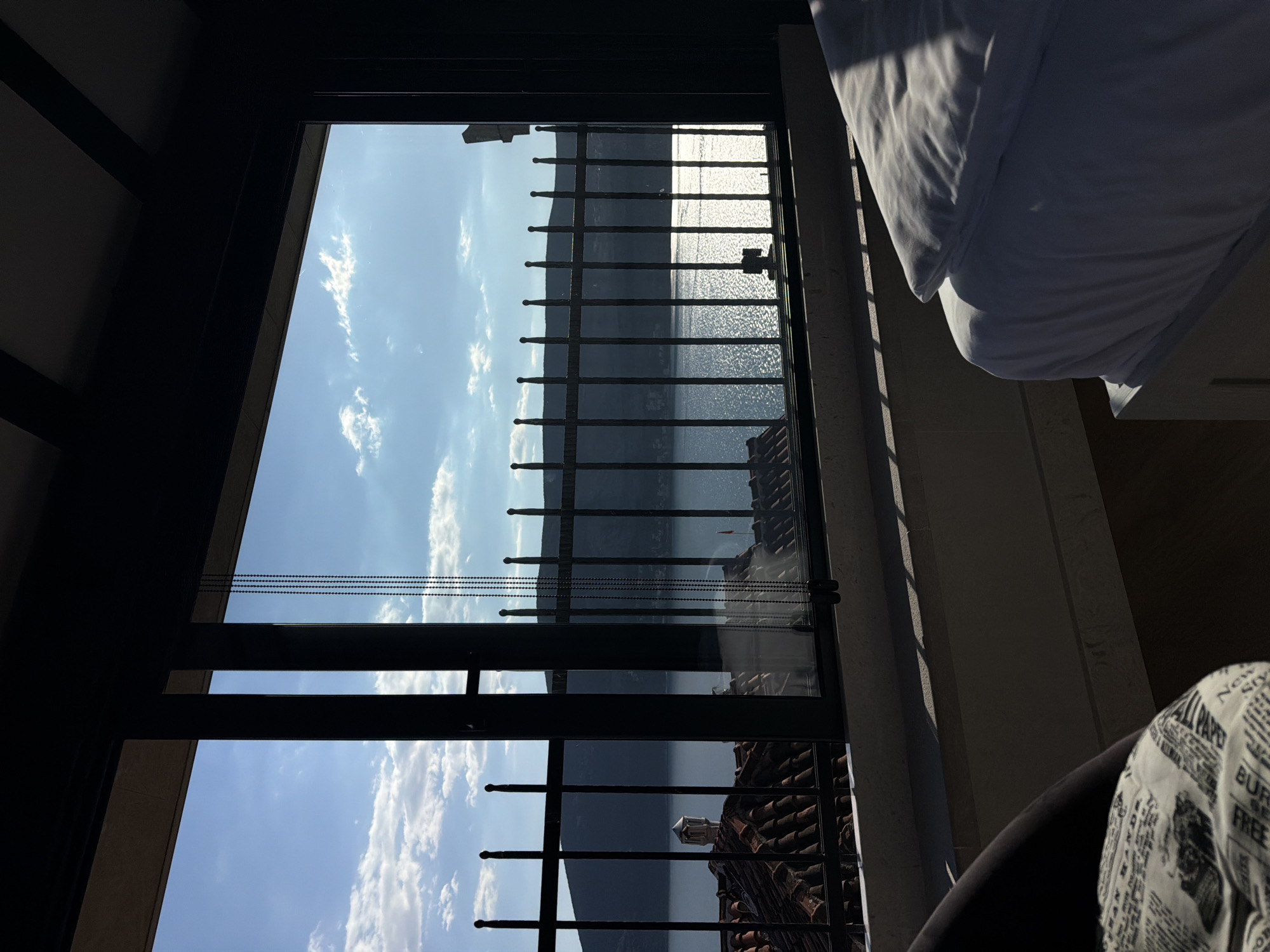 A view from inside a room looking out through a window with metal bars. Beyond the window is a body of water, likely a lake or sea, with mountains in the background. The sky is blue with scattered clouds. The roofs of buildings are visible below the window, suggesting an urban environment. Inside the room, a bed with white sheets is partially visible on the right side, and a chair with a patterned fabric on its back is in the lower right corner. The window frame and parts of the room are in shadow. The location appears to be Trebesin, Montenegro, based on the architecture and surrounding landscape.