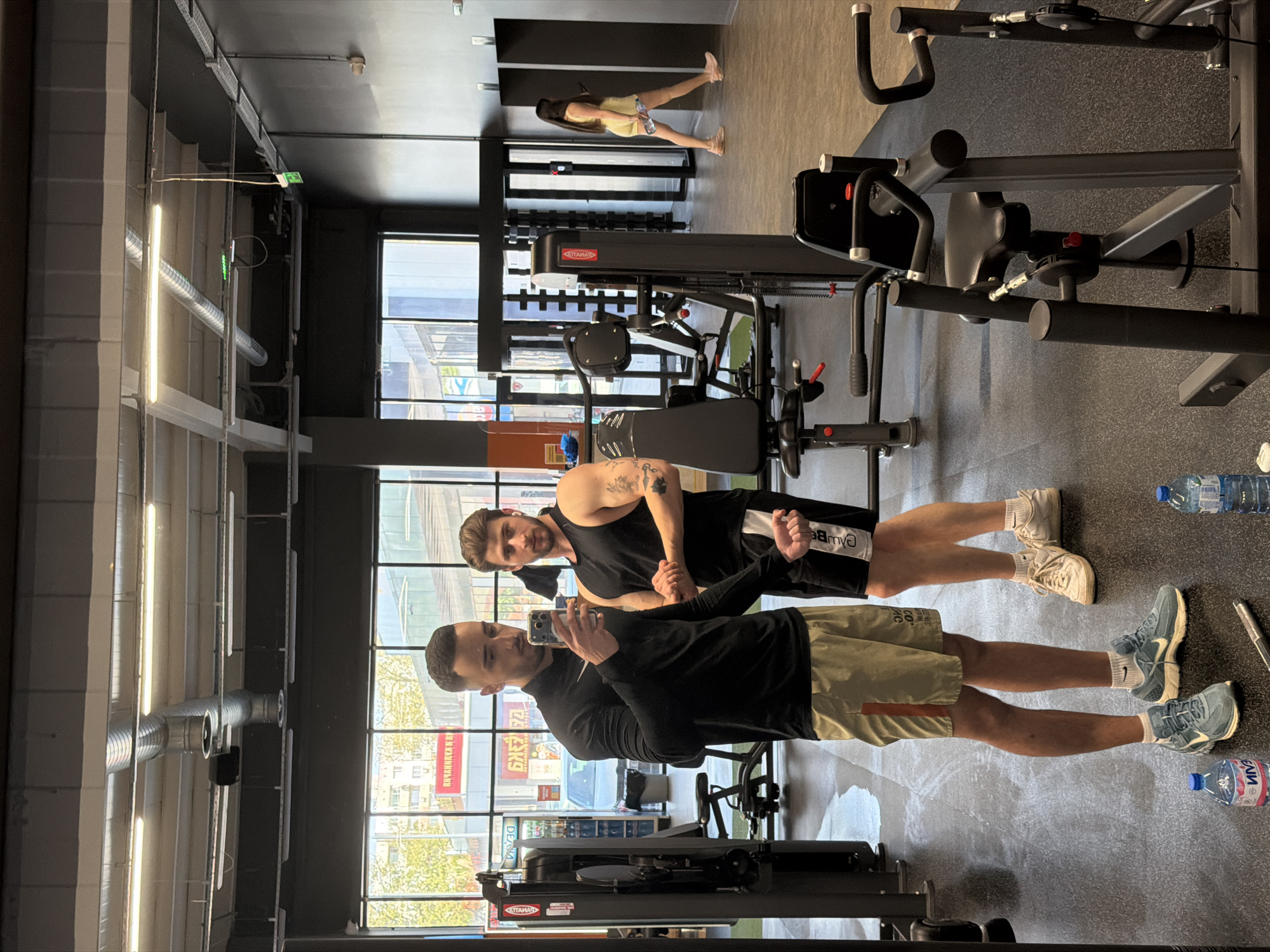This media file captures an indoor scene within a modern gym or fitness center, likely in Plovdiv, Bulgaria, during daytime.
The primary subjects are two men in the foreground, posing for a selfie taken by one of them using a smartphone. The man holding the phone wears a black long-sleeved shirt, olive green shorts, white socks, and light blue sneakers. The man next to him is dressed in a black sleeveless top, black shorts with "Campbe" (possibly part of "Campbell") visible in white text, white socks, and off-white sneakers. He also has tattoos visible on his left arm and shoulder. Both men have short dark hair and beards, and appear casual and content.
In the background, further into the gym, a woman with long dark hair is visible. She is wearing a light yellow top and possibly shorts, and appears to be stretching or standing near other gym equipment. A fourth person's lower body is partially visible in the far background, suggesting more individuals are present.
The setting is characterized by a spacious layout with various gym equipment, including weight machines (one branded "ATAMAH") and what appears to be a rowing machine. Large windows on one wall provide ample natural light, revealing an urban exterior with buildings and some greenery. Through these windows, Cyrillic text can be seen on outdoor signs, such as "РЕКЛАМА" (Advertising), consistent with the provided location context of Plovdiv, Bulgaria.
Architectural details include exposed metallic ventilation ducts and fluorescent tube lighting running across the high ceiling. A green illuminated exit sign is also visible overhead. On the dark gray speckled floor, two plastic water bottles are present, one with "ОМЕГА" (Omega) visible on its label.