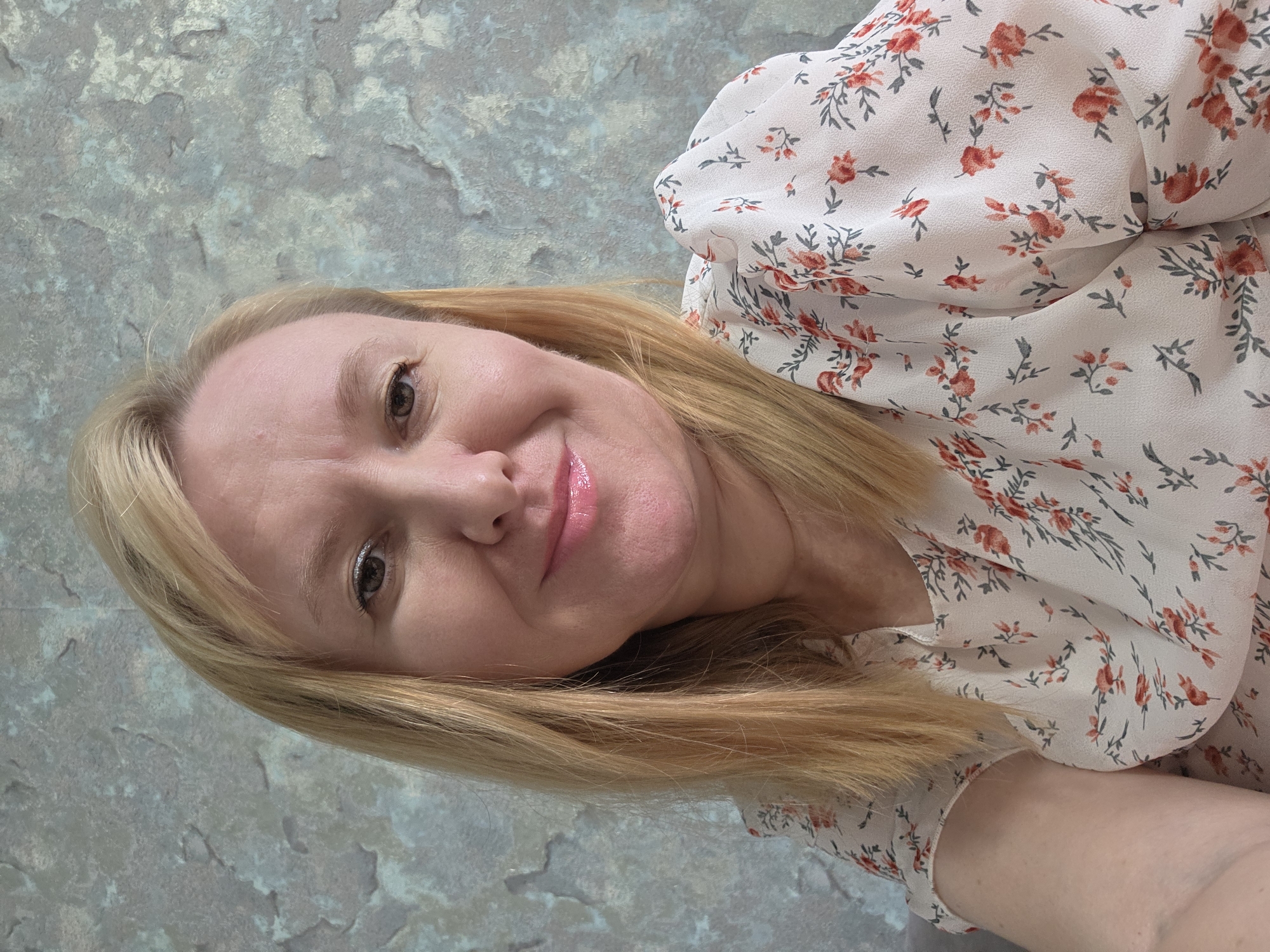 A woman with blonde hair and brown eyes smiles at the camera. She is wearing a white floral-print dress with red flowers and green leaves. The background is a textured gray and blue wall. The image is taken from a close-up perspective, focusing on the woman's face and upper body. The lighting suggests it is daytime, and the setting appears to be indoors. The woman's expression is happy and relaxed.