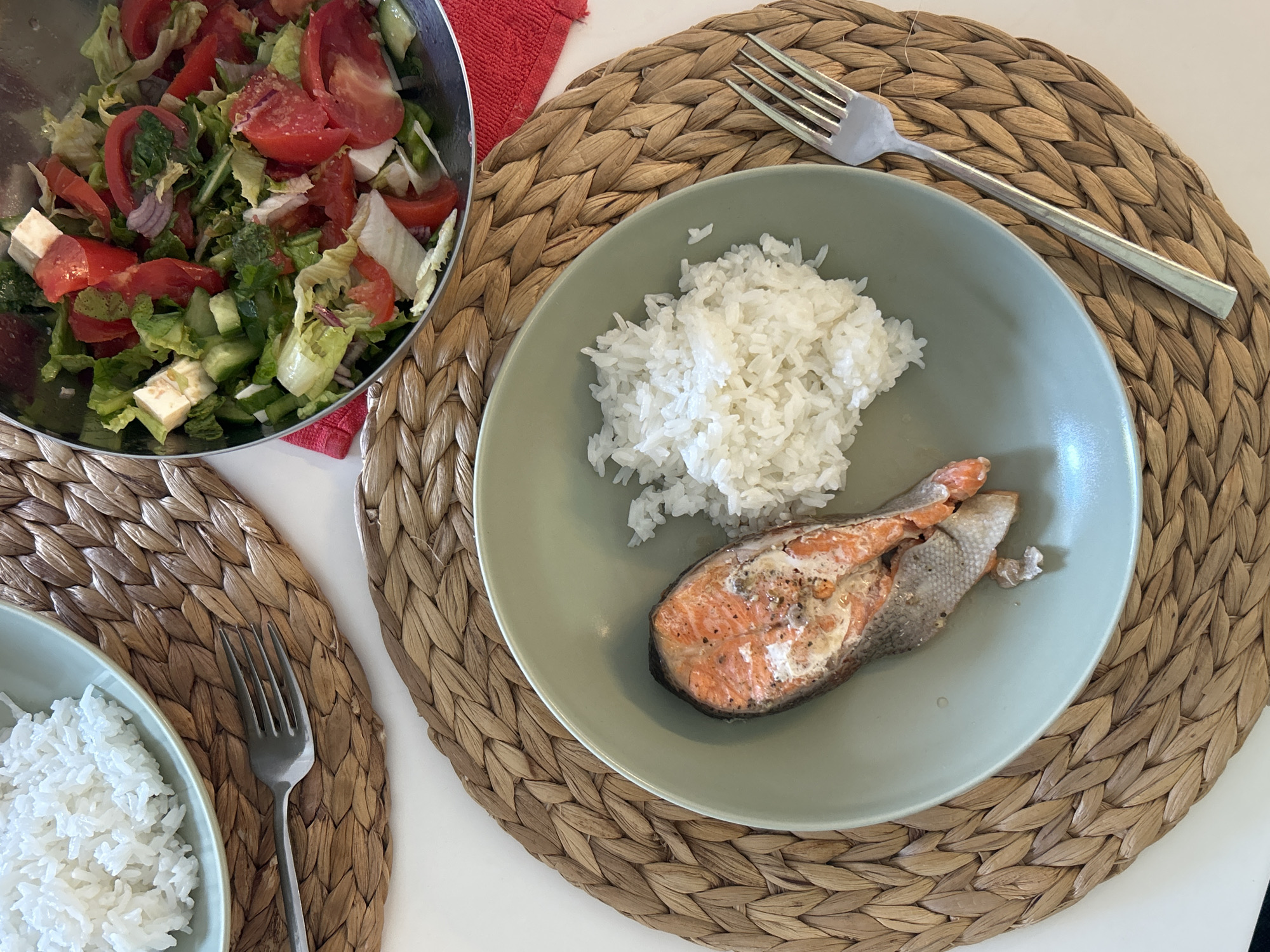 The image shows an overhead shot of a meal served on a woven mat. A light green plate holds a portion of cooked white rice and a piece of salmon. A silver fork rests on the mat next to the plate. To the left, a stainless steel bowl is filled with a salad of lettuce, tomatoes, cucumbers, and feta cheese. Another small green bowl with rice and a fork are visible in the lower-left corner of the image. The overall setting appears to be a dining area, possibly in a home. The location is Alanya, Turkey.