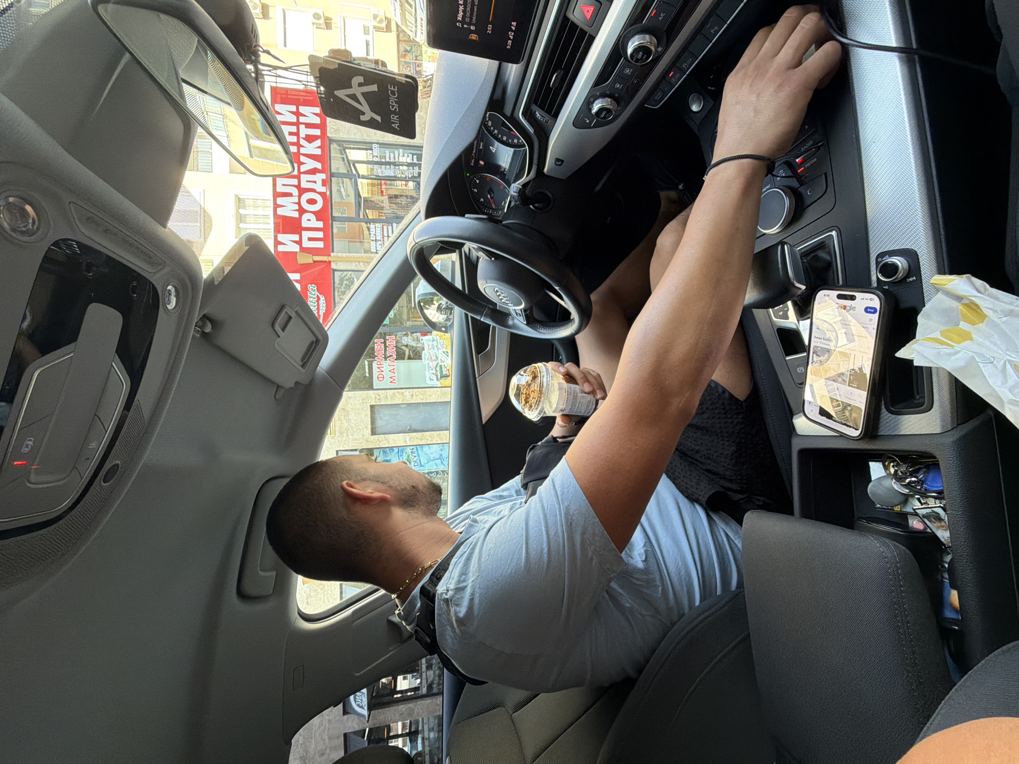 An adult male with a buzz cut and a light blue t-shirt is seated in the driver's seat of a modern car, looking out the passenger window. He is holding a to-go cup with a beverage and ice in his right hand. A black strap is visible on his left shoulder, suggesting a possible injury or brace. The car's interior is visible, including the steering wheel, dashboard with a digital display, center console with various buttons and controls, and a smartphone lying on the console. The phone displays a map interface, indicating navigation is in progress. In the background, through the car windows, a building with a prominent red sign that reads "И МЛЕЧНИ ПРОДУКТИ" (Milk Products) is visible, along with other signage in Cyrillic script. The building appears to be a shop or market in an urban setting. An air freshener with an "A" logo and the text "AIR SPICE" is hanging from the rearview mirror. The overall scene suggests the man is driving or waiting in his car in the city of Burgas, Bulgaria, on a sunny day.