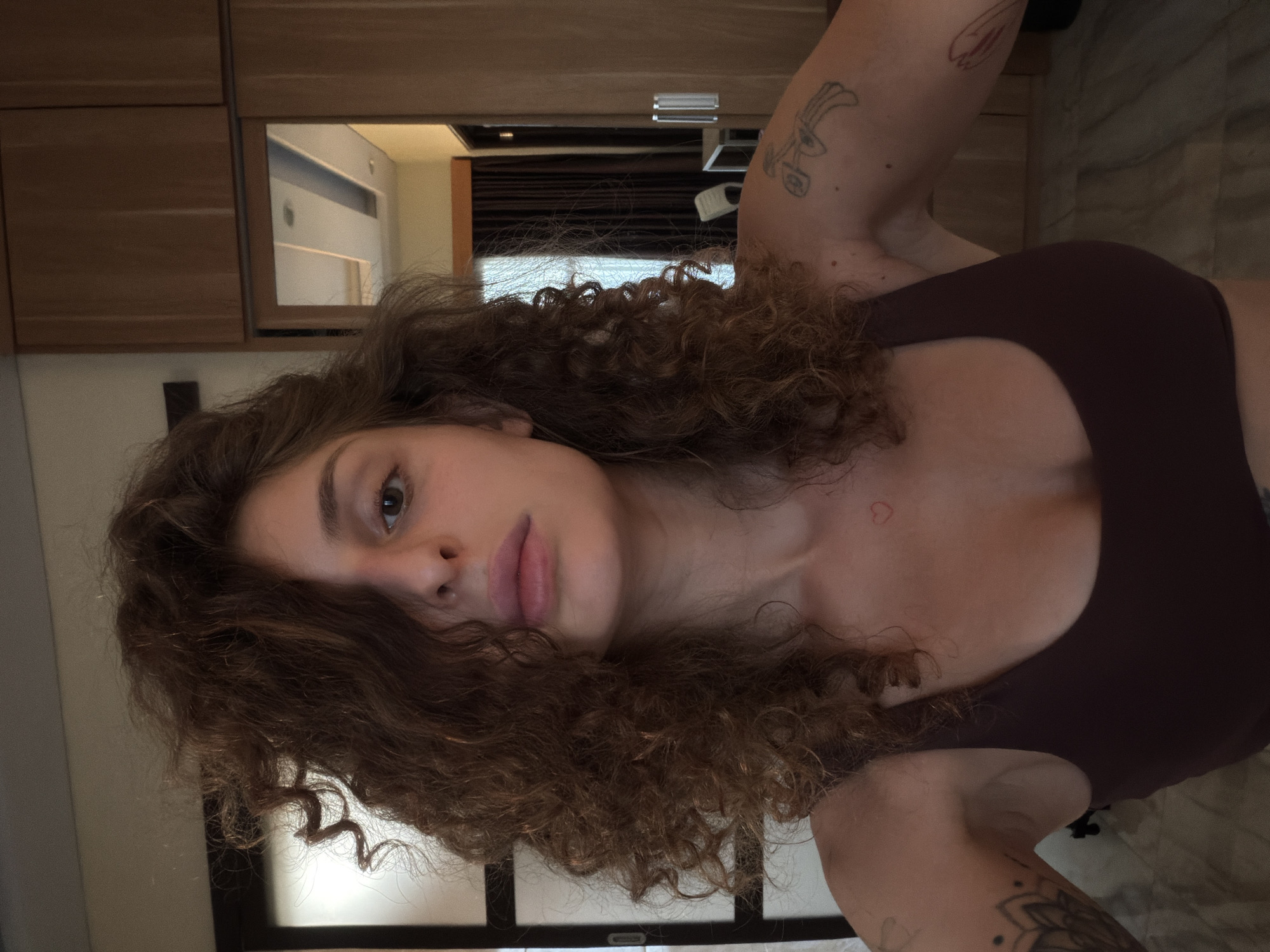 A woman with curly brown hair is facing the camera. She is wearing a dark brown top. She has tattoos on her arms. The background shows a room with wooden cabinets, a mirror, and dark curtains. The location is likely in a hotel or accommodation in North Kuta, Indonesia, based on the interior design.