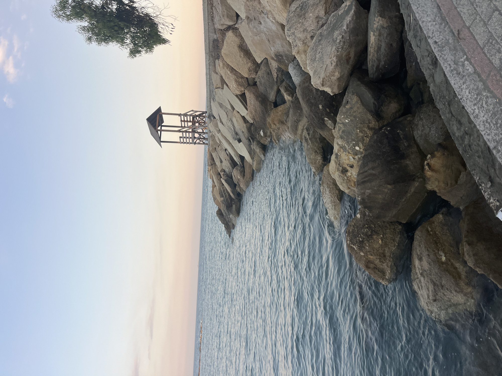 The image shows a rocky coastline in Agios Tychonas Community, Cyprus, during what appears to be sunset or early evening. The sea is calm and slightly rippling. A breakwater made of large rocks lines the shore, with a concrete walkway visible in the foreground. Atop the breakwater, there is a wooden lifeguard tower with a pitched roof and a set of stairs. A tree with green foliage is visible on the right side of the frame, extending into the sky. The sky is a soft gradient of pale blue and pink. No people are visible in the image.
