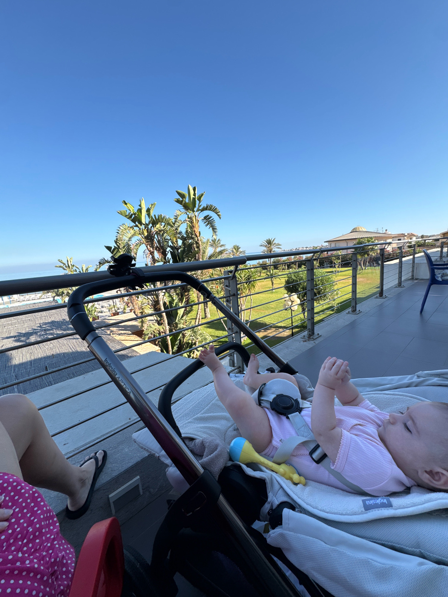 A baby is lying in a stroller on a balcony overlooking a park and the Mediterranean Sea in Capaci, Italy. The baby is wearing a pink onesie and is secured with a harness. A yellow and blue toy is next to the baby. A person's legs are visible on the left side of the image. The balcony has a metal railing and a tiled floor. In the background, there are palm trees, green grass, and buildings. The sky is clear and blue. The sun is shining, casting shadows on the balcony.