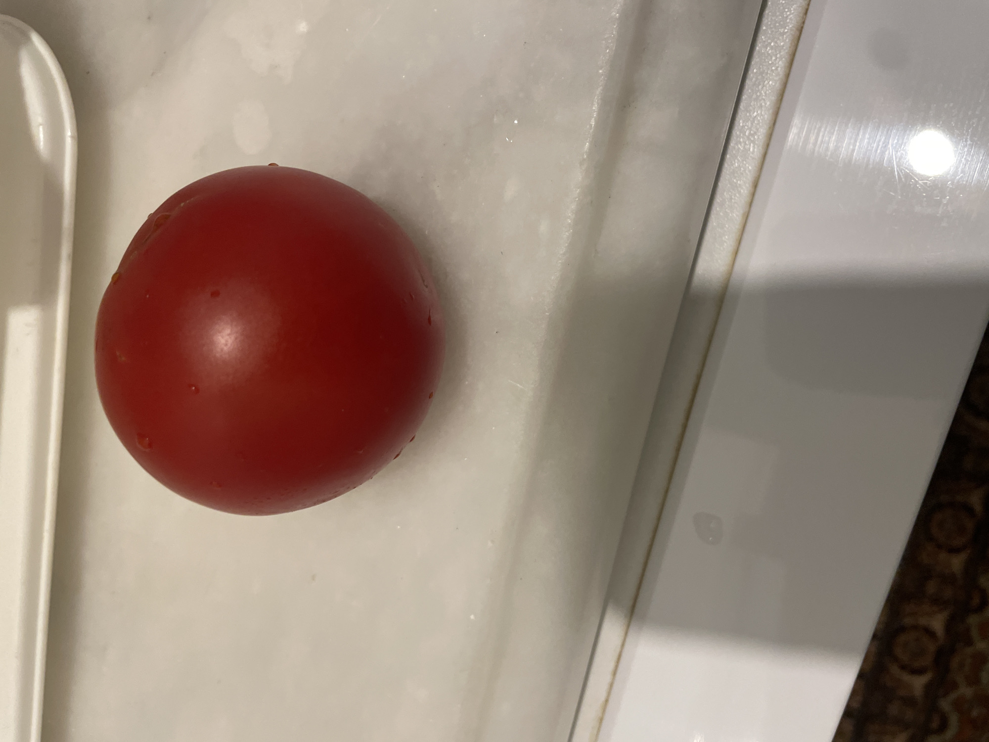 A close-up, eye-level shot shows a single, ripe red tomato resting on a white, slightly textured surface. The tomato is round and appears to be fresh, with small water droplets visible on its skin. The surface it rests on has a slight sheen and is positioned in front of a raised, white border. The lower portion of the image reveals a darker, patterned surface, possibly a rug or tablecloth, and a sliver of a metallic, reflective surface, suggesting the edge of a counter or appliance. The scene is indoors, with neutral lighting. The specific location in Kadıköy, Turkey, cannot be determined from the visual information provided.
