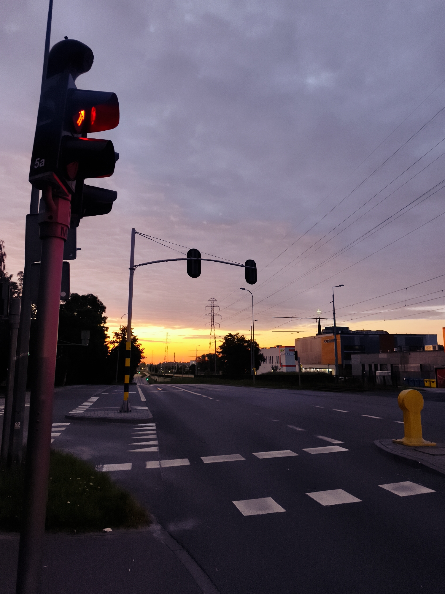 The image captures a street scene at dawn or dusk in Gdańsk, Poland. A traffic light in the foreground is illuminated with a red signal, displaying an arrow pointing upwards and to the left, indicating a directional restriction. The sky is filled with hues of purple, pink, and orange, suggesting the sun is either rising or setting. A road stretches into the distance, with white crosswalk markings and lane dividers. In the background, buildings and power lines are visible. The overall atmosphere is serene and quiet, with no people or vehicles present in the frame. The scene is devoid of any discernable action or interaction, focusing solely on the urban landscape during the twilight hours.