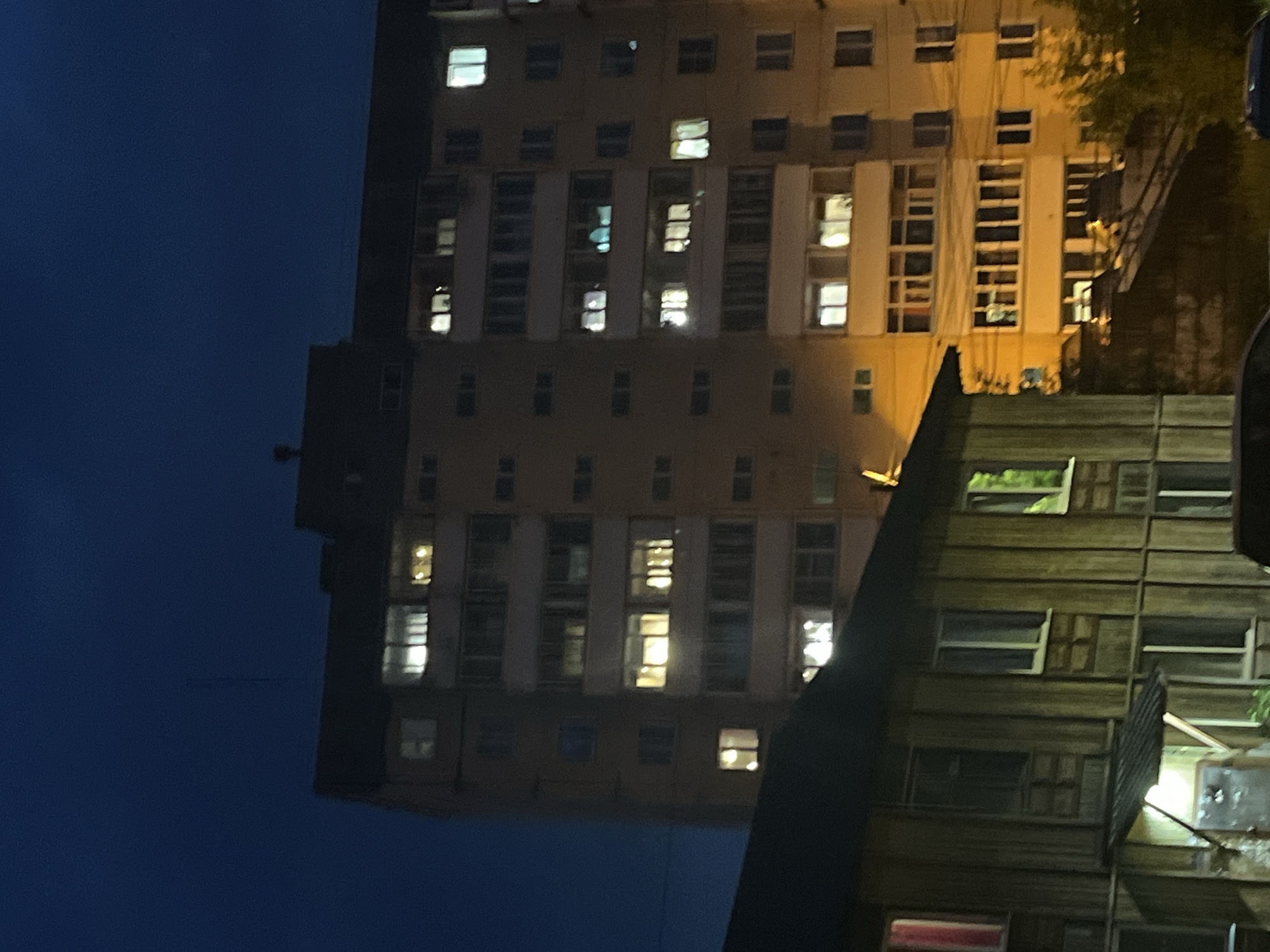 This image depicts a nighttime view of an apartment building and a smaller wooden structure in Divnogorsk, Russia. The sky is a deep blue, indicating late evening or night. The main subject is a multi-story apartment building with numerous illuminated windows, suggesting habitation. Some windows show bright, white light, while others emit a warmer, yellowish glow. The apartment building has a dark roofline and appears to be made of concrete or a similar material.
In the foreground, partially obscuring the lower portion of the apartment building, is a smaller, two-story wooden structure. This building has vertical wooden siding and several windows, some of which are also lit, though less brightly than those in the apartment building. A small, covered entranceway with a light is visible on the left side of the wooden structure.
The overall scene suggests a residential area at night. The lighting from the windows creates a contrast against the dark sky. The presence of both a large apartment building and a smaller, older-looking wooden structure indicates a diverse urban landscape. The image captures a moment of quiet domestic life within the city.