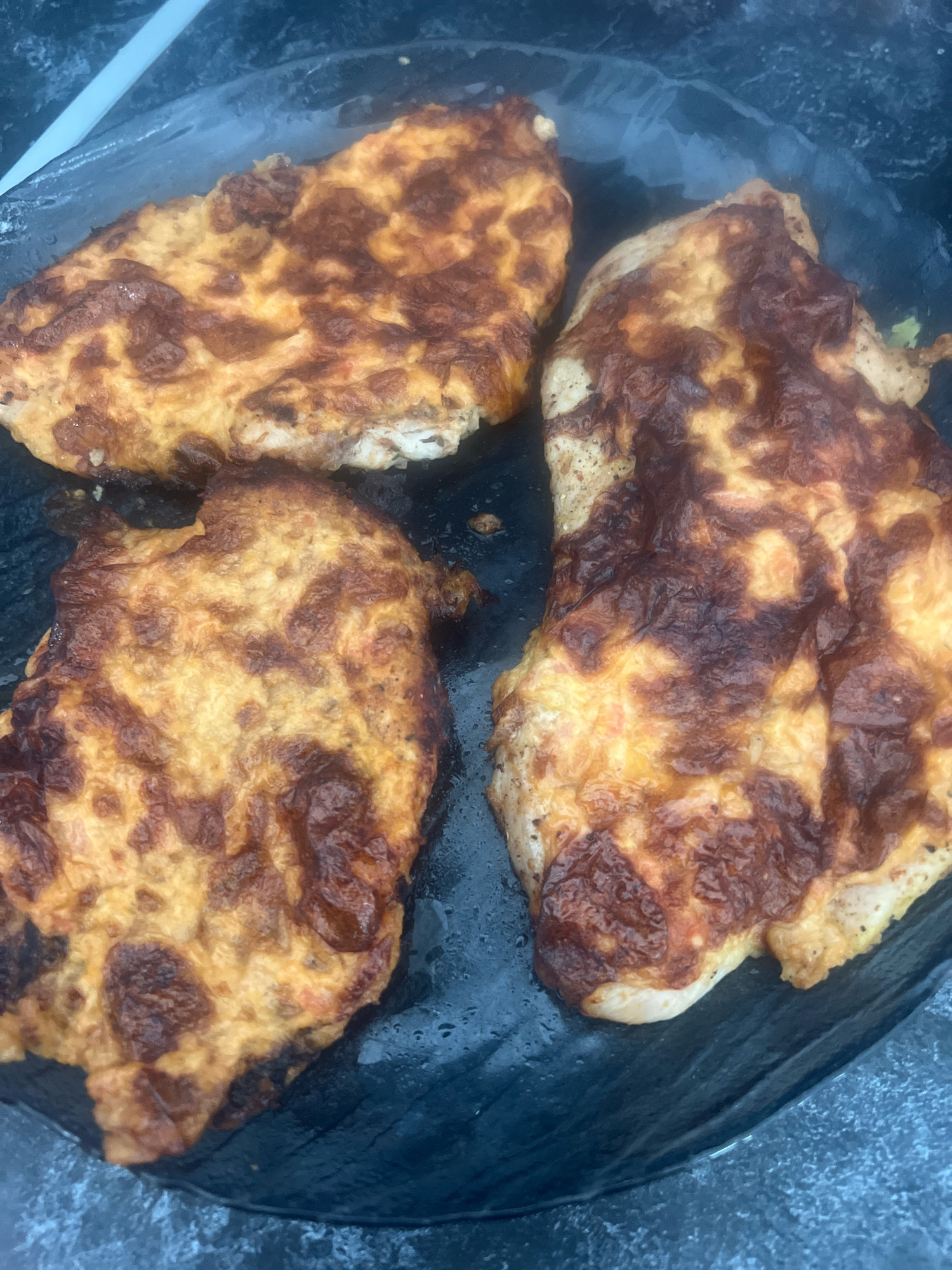 Three baked chicken cutlets are presented on a dark, wet surface, possibly a platter or a countertop. The chicken cutlets are covered in a melted, golden-brown cheese topping with some areas appearing slightly charred, indicating they have been baked or broiled. The surface beneath the cutlets has a textured, dark appearance and is visibly wet, with small droplets and streaks of moisture. The lighting in the scene is somewhat dim, casting shadows and highlighting the sheen of the wet surface and the texture of the cooked cheese. There are no people visible in the image, and the focus is solely on the food. The scene does not provide any specific visual cues to identify its location within Otradnoye, Russia, or any other city.
