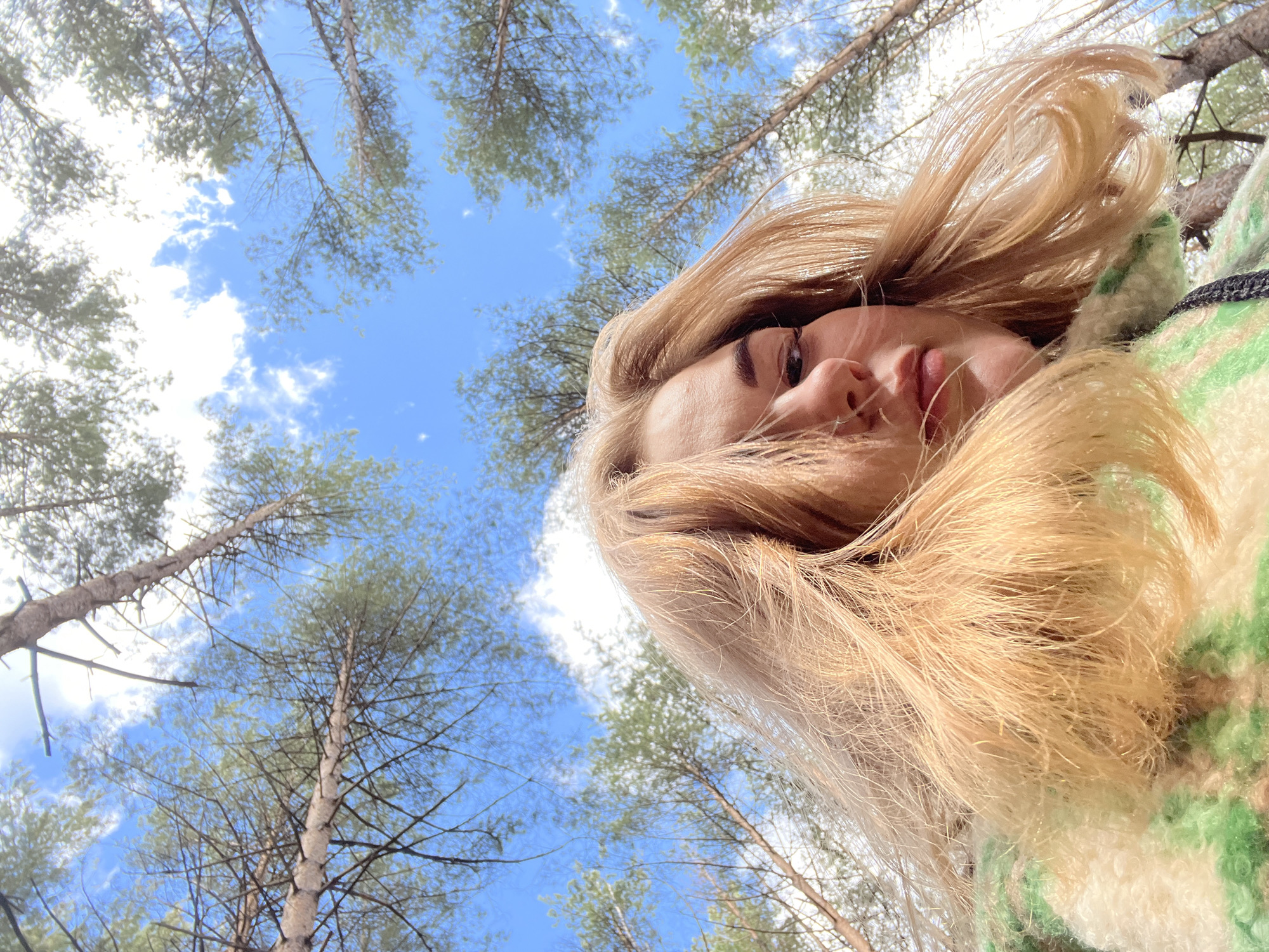 A woman with blonde hair is lying on her back in a forest, looking up at the sky. The trees are tall and slender, with green foliage. The sky is blue with white clouds. She is wearing a green and white sweater. The location is possibly Chaikovsky, Russia, given the context of the prompt.