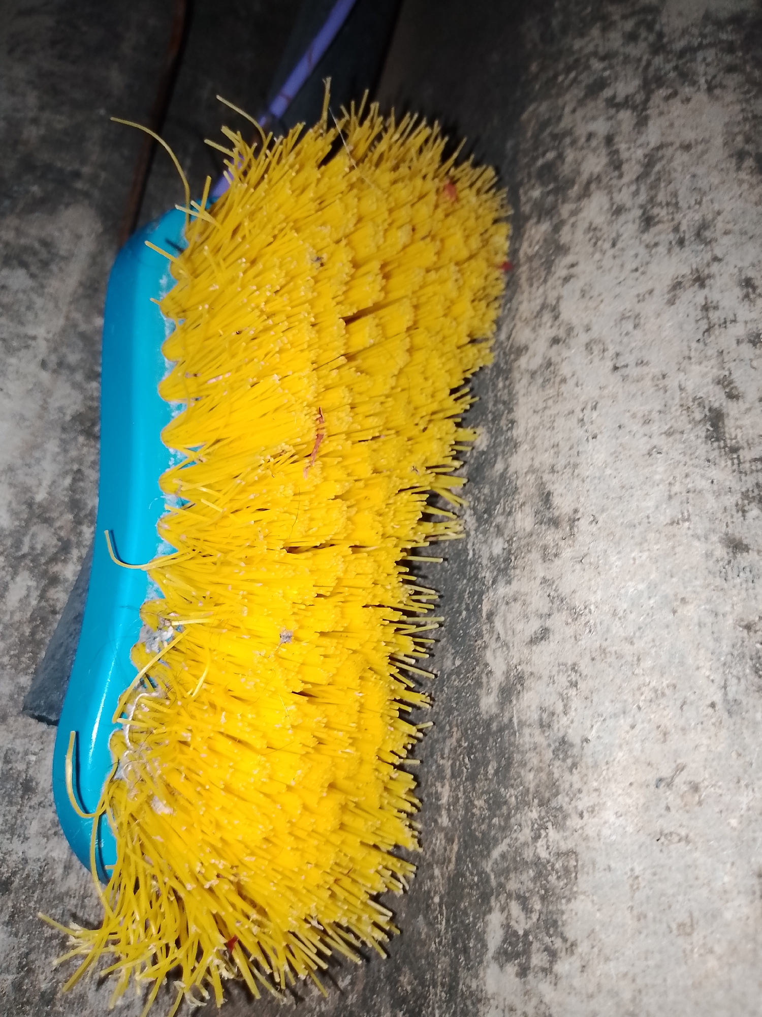 A close-up, overhead, eye-level shot captures a bright yellow brush with a blue handle resting on a rough, gray concrete surface. The brush bristles are densely packed and appear to have some white and reddish debris clinging to them. A few stray bristles curl outwards from the main cluster. In the background, a darker, textured surface with horizontal lines is visible, possibly indicating wooden planks or a similar material. The scene is dimly lit, casting faint shadows. The image was taken in Santa Ana, Venezuela.