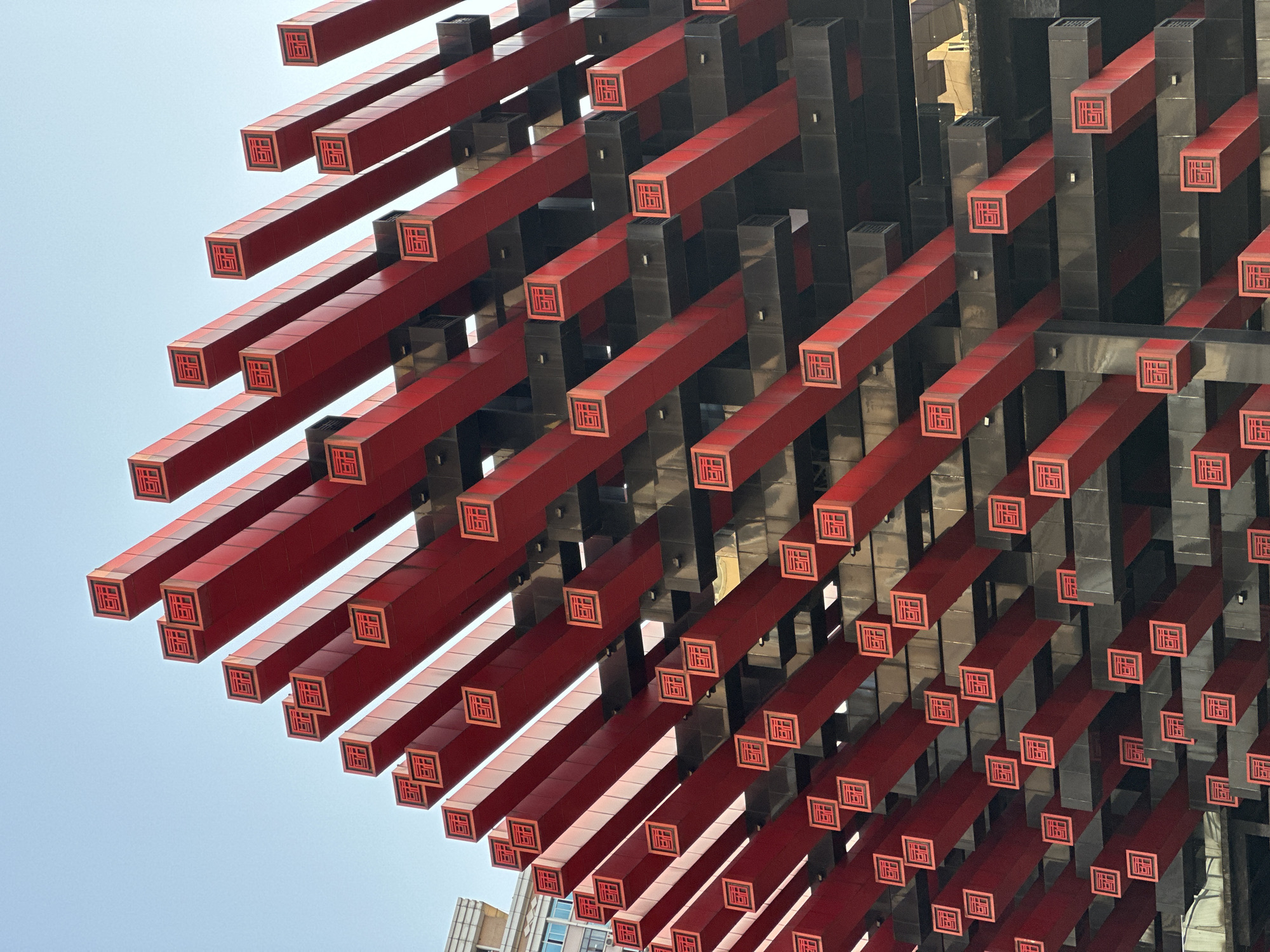 The image shows a portion of the Raffles City Chongqing building, located in Yuzhong District, China. The view is angled upwards towards the structure, which features a repetitive pattern of red and black rectangular beams protruding outward. Each red beam has a square imprint with a design on its end. The sky is visible in the upper left corner. A building is visible in the lower left.