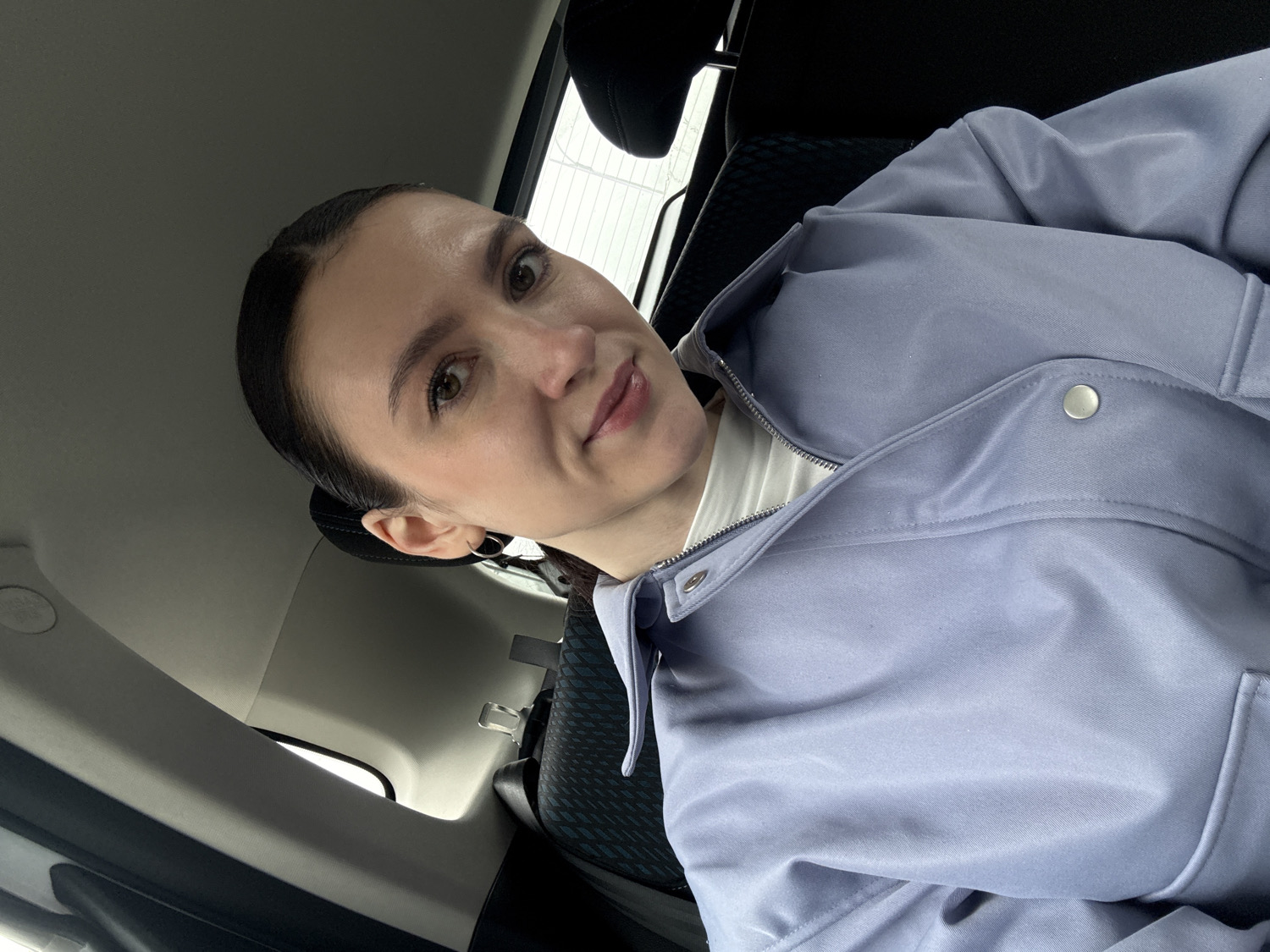 A woman is depicted in a selfie-style photograph taken inside a car. She is positioned towards the right side of the frame, looking directly at the camera with a slight, natural smile. Her dark hair is pulled back from her face, revealing her ears, one of which features a dark earbud or earring. She has brown eyes and appears to be wearing minimal makeup.
She is dressed in a light, muted purple or lavender jacket, which is partially unzipped, revealing a white top underneath. The jacket features silver snap buttons along the placket. The car's interior is visible behind her, characterized by dark-colored upholstery on the seats and headrests. A window in the background allows natural light to enter, suggesting it is daytime, possibly overcast, or she is in an indoor parking structure.
Faint text is visible on the car's ceiling or pillar in the upper left portion of the image, showing "AIRBAG" and "SRS" below it, indicating a safety feature. The overall impression is a casual moment captured within a vehicle.