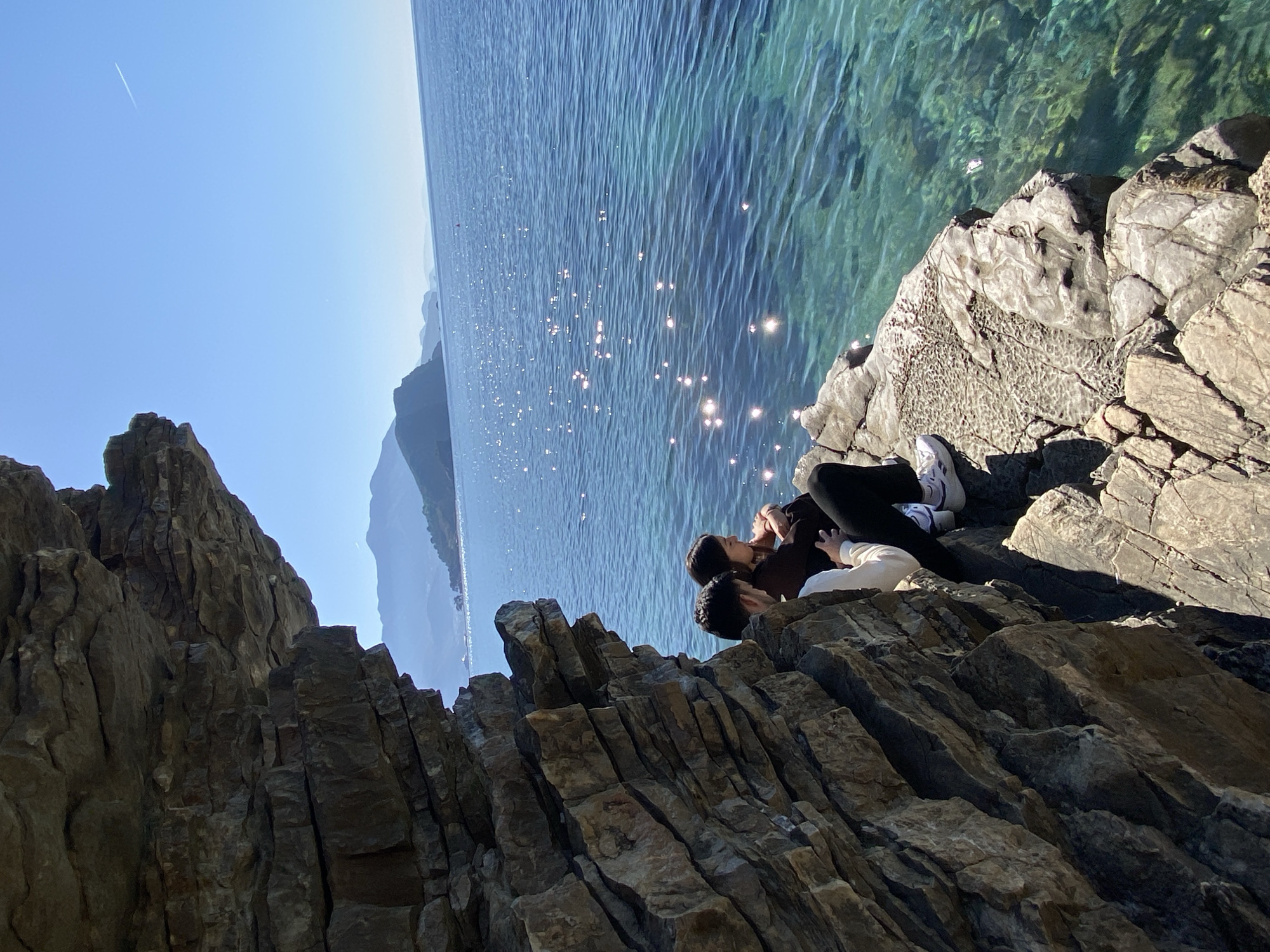 A couple is lying on a rocky outcrop overlooking the Adriatic Sea near Budva, Montenegro. The sea is blue with sparkling sunlight. The mountains are in the background. The sky is clear. The rocky outcrop is dark and striated. The couple are lying close to each other. The person on the left has dark hair, is wearing a brown top, and black trousers. The person on the right has dark hair, is wearing a cream top, and black trousers with white shoes. The location is likely along the coastal area near Budva, known for its rocky beaches and clear waters.
