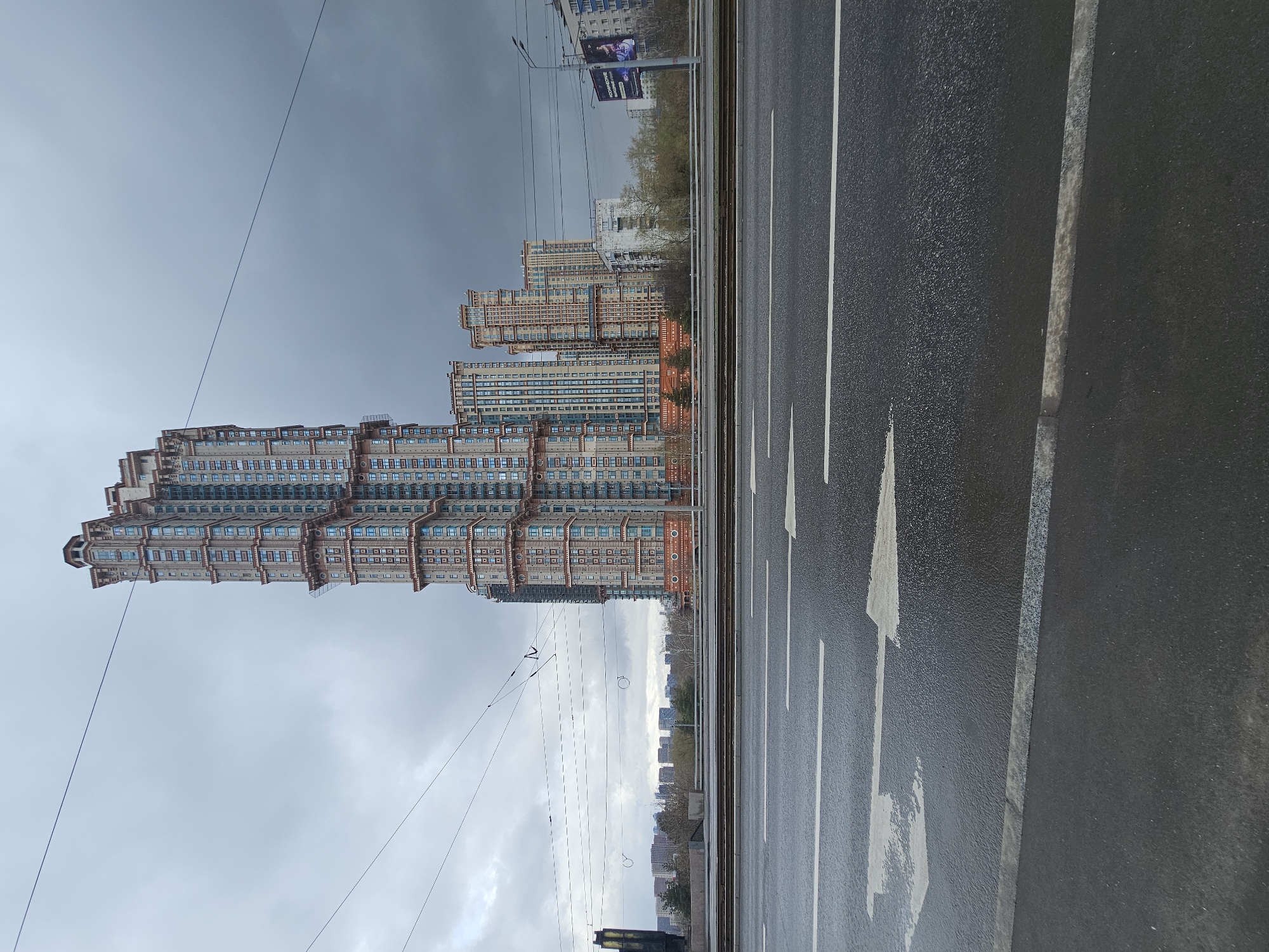 The image shows a street scene in Khimki, Russia, under an overcast sky. The foreground is dominated by an asphalt road with white lane markings and a large arrow indicating the direction of traffic. To the left of the road are high-rise buildings with a distinctive architectural style. The buildings have a layered appearance with a mix of brick and blue glass. Tram lines and overhead wires are visible, adding to the urban setting. In the distance, more buildings are seen, suggesting a densely populated area. The overall environment is urban and possibly industrial.