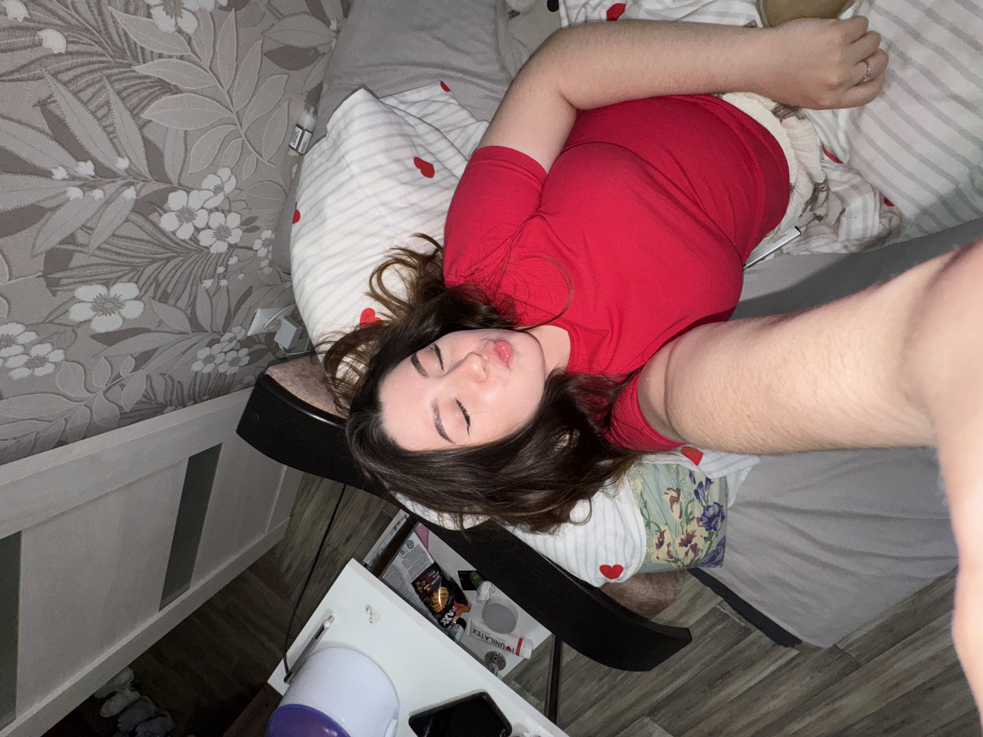 An overhead selfie captures a young woman lying on a bed in what appears to be a bedroom. Her head is towards the bottom of the frame, with her body extending diagonally upwards. She has dark hair, closed eyes, and her lips are pursed in a playful or relaxed expression. She is wearing a red t-shirt. Her right arm is stretched towards the top of the frame, likely holding the camera, while her left arm is bent near her head.
The bed features a gray headboard and a light-colored wooden frame. The bedding includes pillows with white and gray stripes adorned with small red hearts, and a floral patterned blanket or duvet in green, purple, and white. A plain gray sheet is also visible. The adjacent wall is covered with light gray wallpaper featuring a large pattern of white and darker gray leaves and flowers.
To the lower left, a white nightstand or small table is seen. On it rests a white and purple cup or container, a black smartphone, and a silver pen. A partially open drawer reveals various items, including a snack bag with orange/yellow packaging (partially showing "max") and a white box labeled "UNILATEX". Underneath the bed, faint outlines of what might be shoes or slippers are visible on the greyish wooden floor, which is laid in a patterned style. A small bottle with text like "VANILLA CANDY" is near the wall.
The scene suggests a relaxed, indoor setting, likely a personal moment captured in a bedroom. There are no strong cues about the time of day or weather beyond it being indoors. The mood conveyed by the subject's expression is one of calm or lightheartedness. The location context is Savenki, Russia.