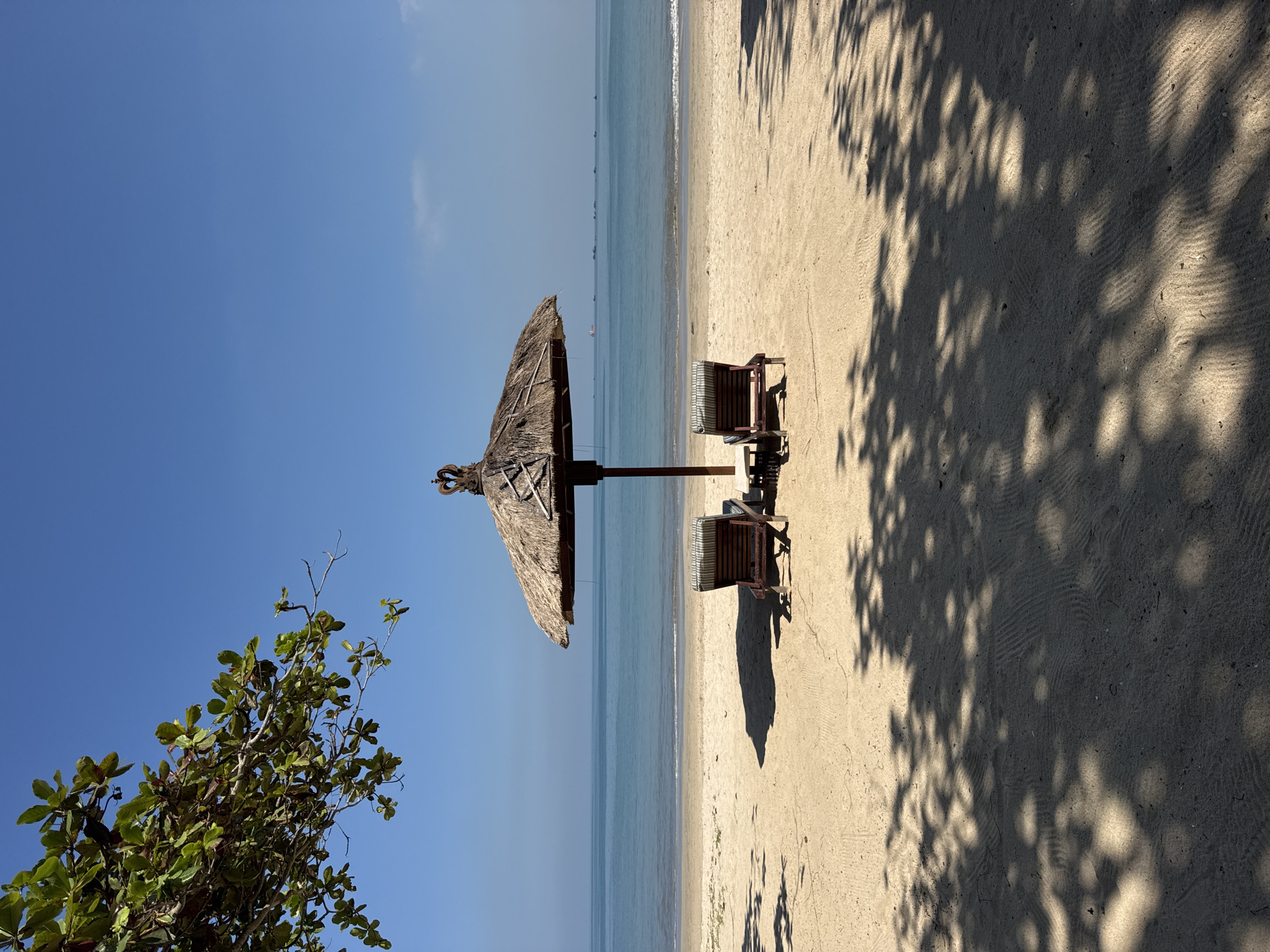 This image captures a tranquil tropical beach scene, likely in Kuta Selatan, Indonesia, under a bright, clear blue sky.
In the midground, a prominent thatched umbrella (palapa) provides shade over two empty wooden beach chairs, which are positioned on the light-colored sand. The chairs feature striped cushions and face towards the ocean. The strong overhead sunlight casts distinct, dappled shadows from trees and the umbrella across the sandy beach, particularly on the right side of the frame, indicating either mid-morning or late afternoon. The sand itself shows subtle textures and ripples.
To the left, green foliage from a tree partially frames the scene against the vibrant blue sky, suggesting a lush, natural environment. Beyond the sandy beach, the calm, blue-green ocean stretches horizontally across the image, meeting the horizon with the clear, unblemished sky. Numerous small boats are visible as faint, dark silhouettes scattered across the distant ocean surface.
The weather is bright and sunny with very few, wispy clouds. The overall atmosphere is one of peace and relaxation, typical of a vacation destination. There is no visible text in the image.