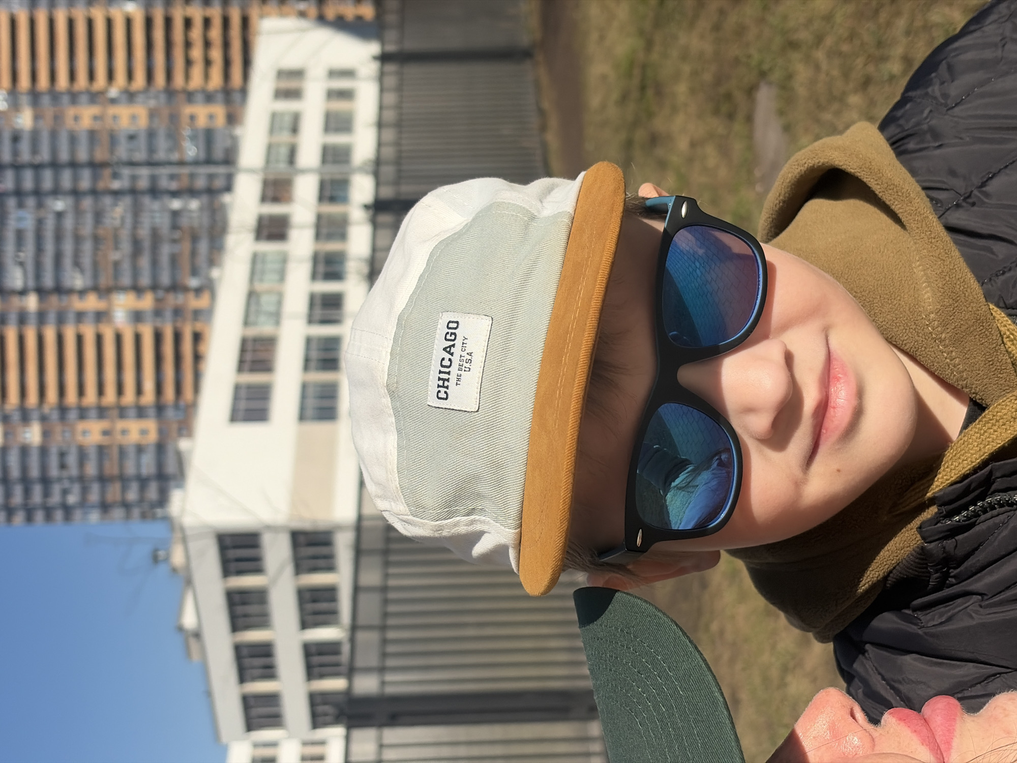 The image depicts a boy wearing sunglasses and a hat with the word "Chicago" on it. He is wearing a tan jacket and a black coat. The background includes buildings and a fence, suggestive of an urban setting. Based on the architecture, it is plausible that the location could be Saint Petersburg, Russia.
