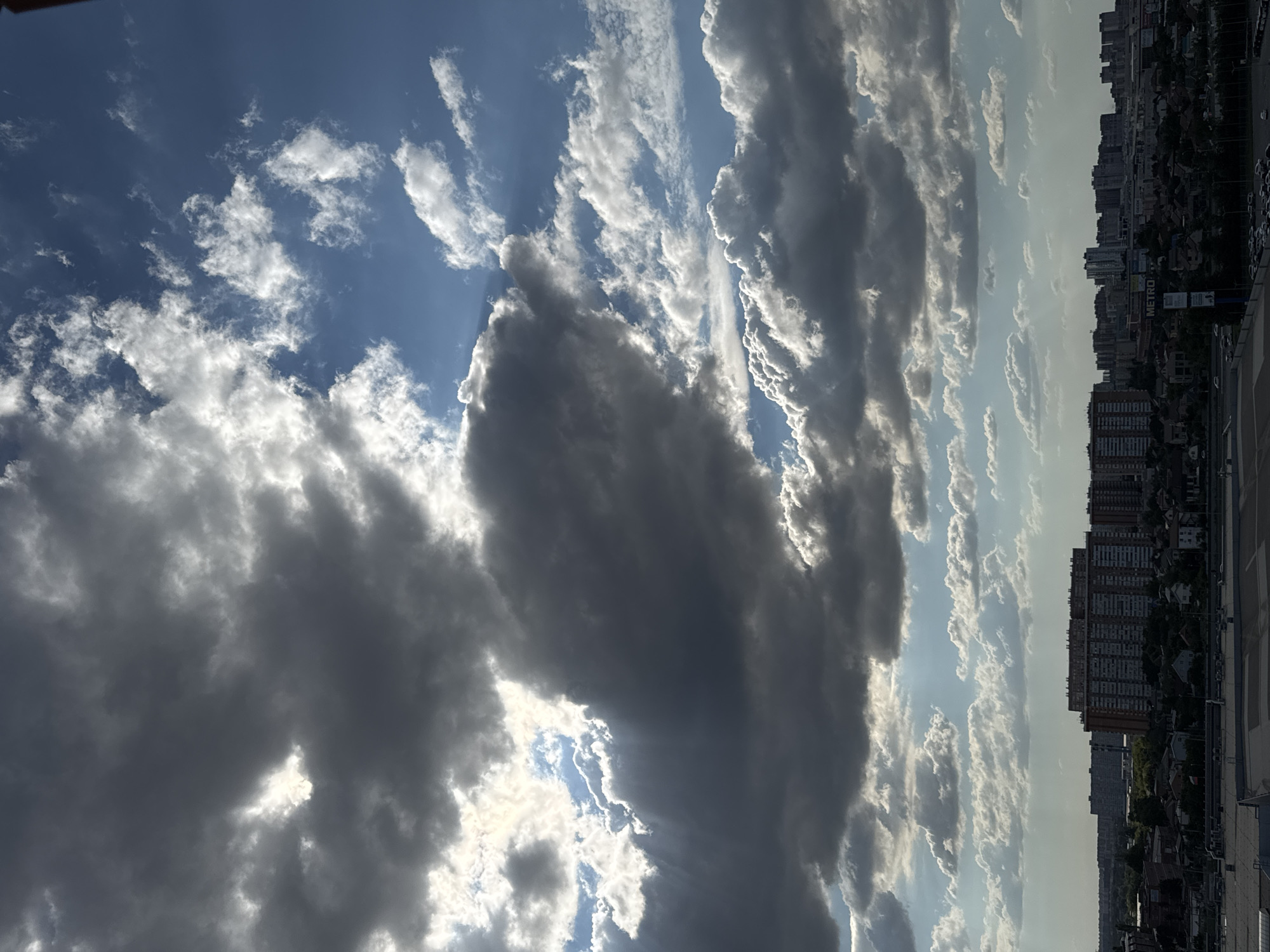 The image depicts a sky filled with dramatic clouds over a city skyline. The clouds are a mix of dark, heavy formations and lighter, scattered cumulus clouds, with sunlight breaking through in some areas, creating visible rays. Below the sky, a cityscape is visible, characterized by numerous apartment buildings and other structures. In the foreground, a large, flat-roofed building, likely a commercial or industrial structure, is present. The overall impression is that of a dynamic sky above an urban environment. The presence of buildings and infrastructure suggests a densely populated area. There are no visible people or specific actions taking place in the image. The lighting indicates it is daytime, with the sun partially obscured by clouds.