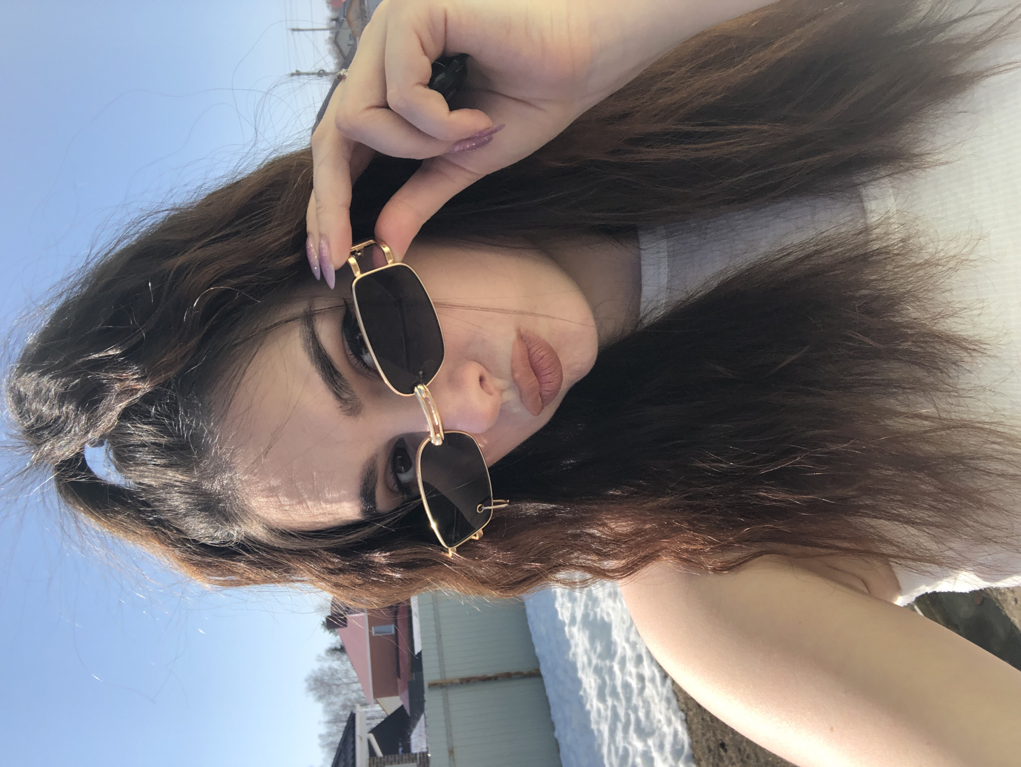 A young woman with long brown hair holds a pair of gold-framed sunglasses in front of her face. She wears a white top. The background shows a clear blue sky, buildings, and snow-covered ground, suggesting an outdoor winter setting. The buildings are small, possibly houses, with a reddish roof visible. The location is identified as Noviy, Russia.