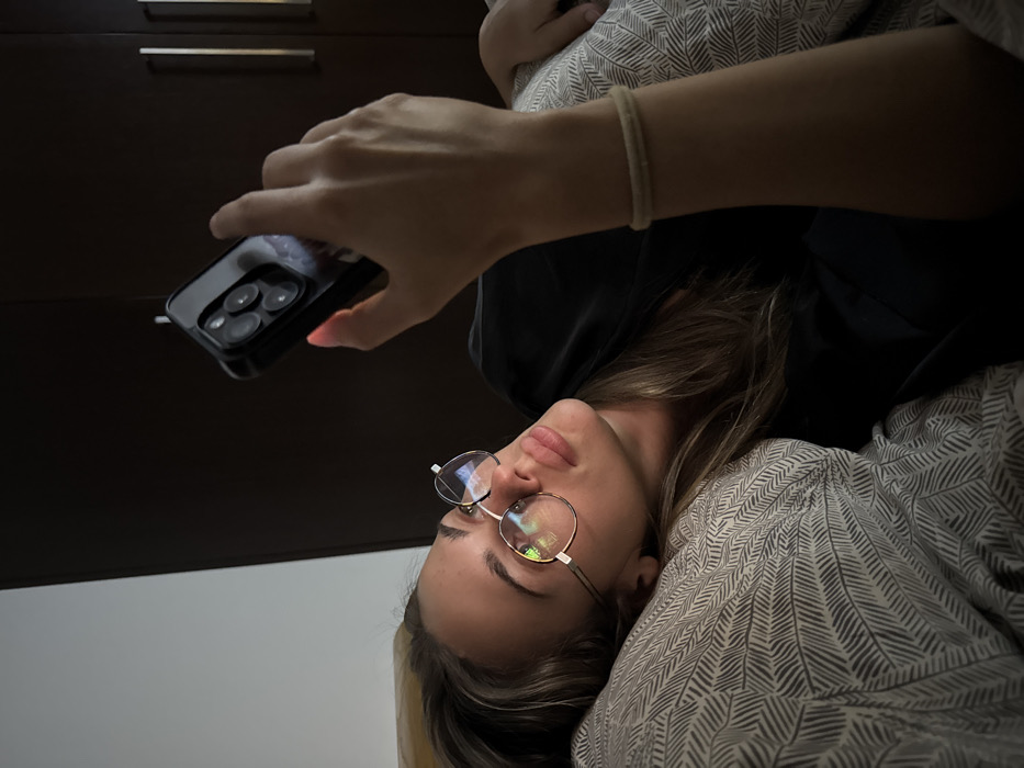 A woman lies on a patterned pillow, gazing upwards at a smartphone held in her outstretched hand. She is wearing round eyeglasses. Behind her is a dark wooden cabinet, and she has a simple bracelet on her wrist. The scene is dimly lit, creating a sense of relaxed intimacy. The angle of the shot is from the side and below, accentuating the upward gaze and the phone's presence.
