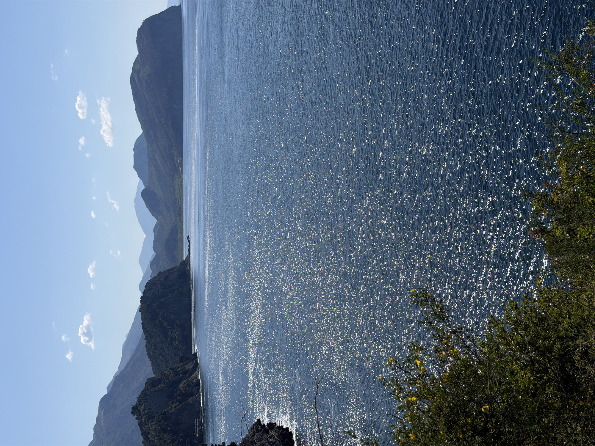 The image shows a landscape view of a lake bordered by mountains. The lake is a deep blue with shimmering sunlight reflecting off the surface. The mountains rise sharply along the left side of the image, fading into the distance with a gradient of blues and greens. In the foreground on the right, there is some green vegetation. The sky is clear and light blue. The location is likely near Petrovac centar, Montenegro, given the mountainous terrain and coastal presence.