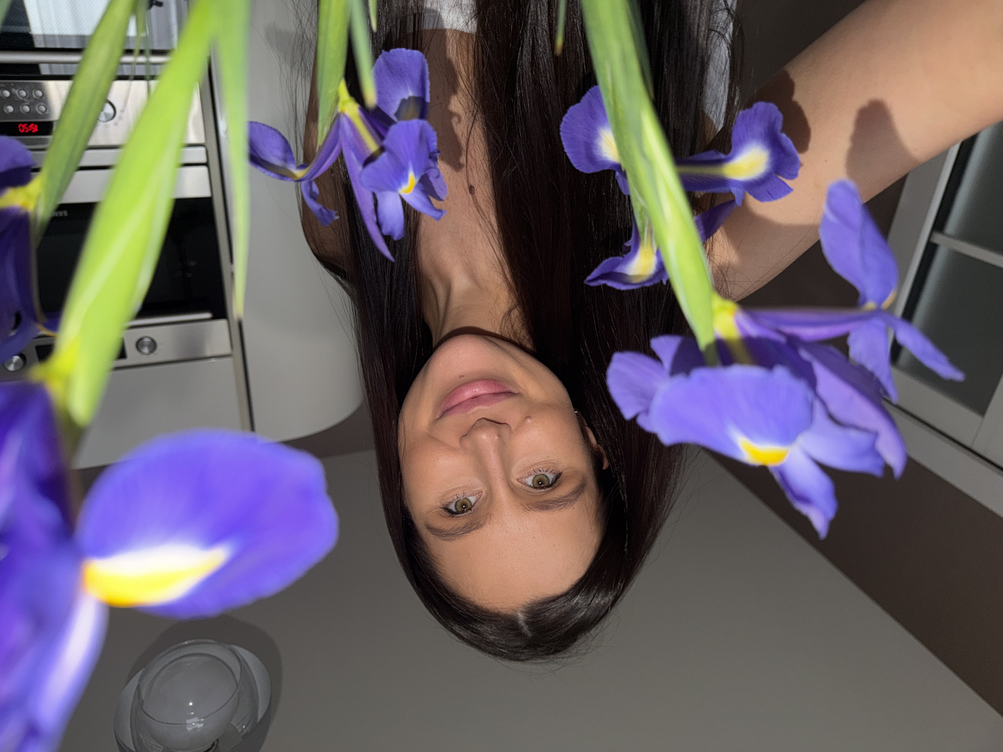 A woman with long, dark hair is looking at the camera. She is positioned in the center of the frame, upside down. Her eyes are hazel, and her skin tone is fair. The background is a modern kitchen with gray walls and white cabinets. In the foreground, there are several violet irises with yellow centers. To the left, a clear vase can be seen. In the background, a microwave oven with a digital display showing "05:59" is visible. The scene's setting is likely within a home. The image was likely taken in Khimki, Russia, or at least portrays a scene consistent with that location.