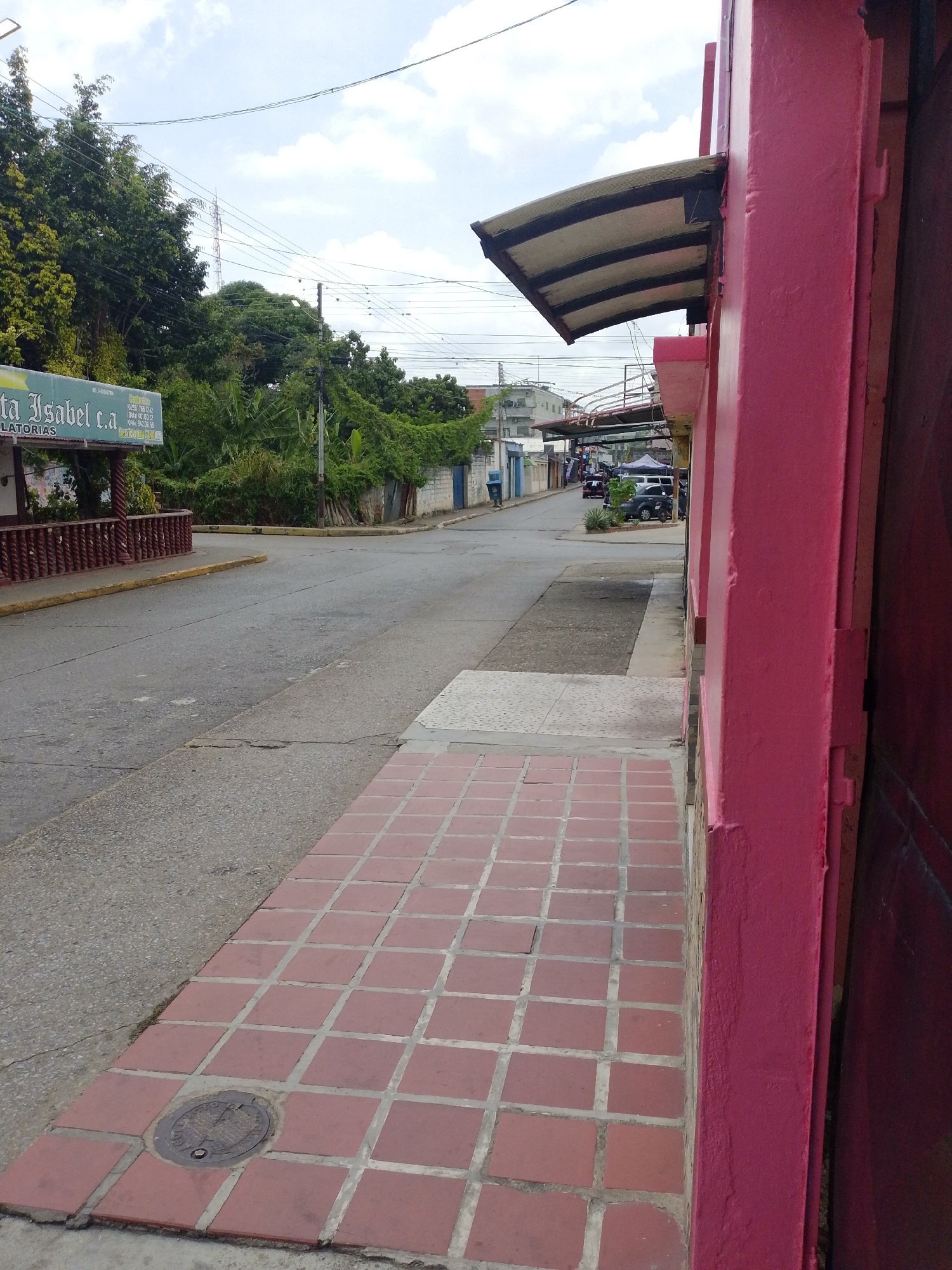The image captures a street scene in Tinaquillo, Venezuela. The viewpoint is from a sidewalk paved with red square tiles, bordering a building painted in a vibrant pink hue. The street extends into the distance, featuring a mix of asphalt and concrete surfaces. On the left side of the street, dense green vegetation lines the road, with a sign for "Santa Isabel c.a" visible. In the middle distance, cars are parked along the street, and pedestrians are seen under a metal structure spanning across the road. The sky is partly cloudy, indicating daytime. Overhead wires are visible running across the scene. A manhole cover is visible in the foreground of the sidewalk.