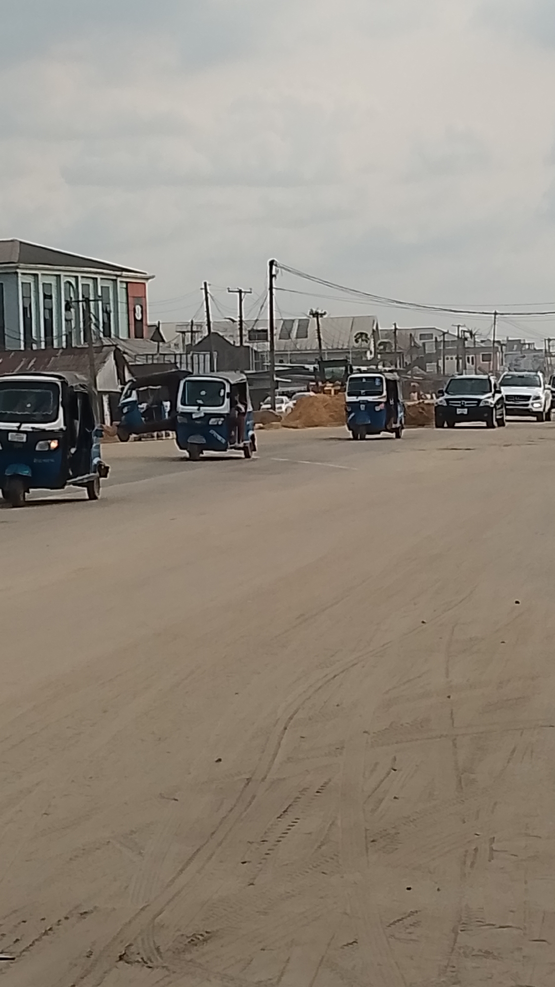 The image depicts a wide, unpaved road in Asubi, Nigeria, likely a peripheral urban area due to the presence of basic infrastructure and mixed building types. The road surface is sandy and marked with numerous tire tracks. Traffic includes three-wheeled motorized vehicles ("Keke Napep") in blue and white, along with two SUVs in the distance. The background features a mix of low-rise buildings, some appearing more established and others temporary or under construction. Utility poles with overhead wires are visible, indicating basic services. The sky is overcast, suggesting the weather is cloudy or dull. No pedestrians are clearly visible, but occupants are present in the Keke Napeps. There are no apparent interactions other than vehicular traffic.