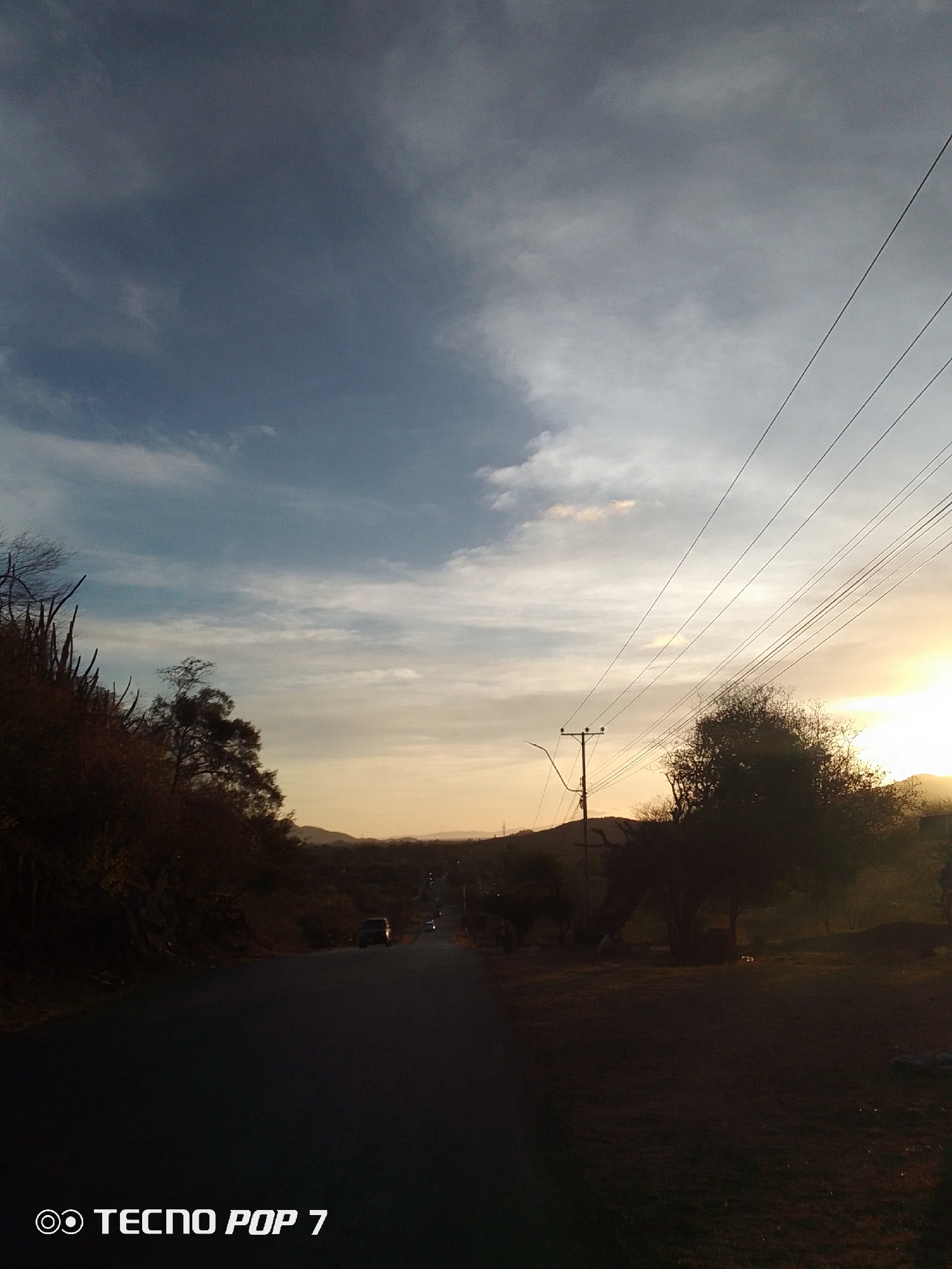The image depicts a road leading into the distance under a partly cloudy sky. The sky is a mix of blue and grey with a band of orange/yellow on the horizon suggestive of either sunrise or sunset. Power lines run along the right side of the road, parallel to the roadway. A utility pole is visible on the right side of the road. The road itself is dark and winding, with some sparse vegetation on either side. At least two vehicles are visible in the distance. The foreground is mostly dark, with a smartphone branding "TECNO POP 7" visible at the bottom. The location is identified as Altagracia, Venezuela, though specific markers of the city are not visible in the image. There are no people visible in the image.