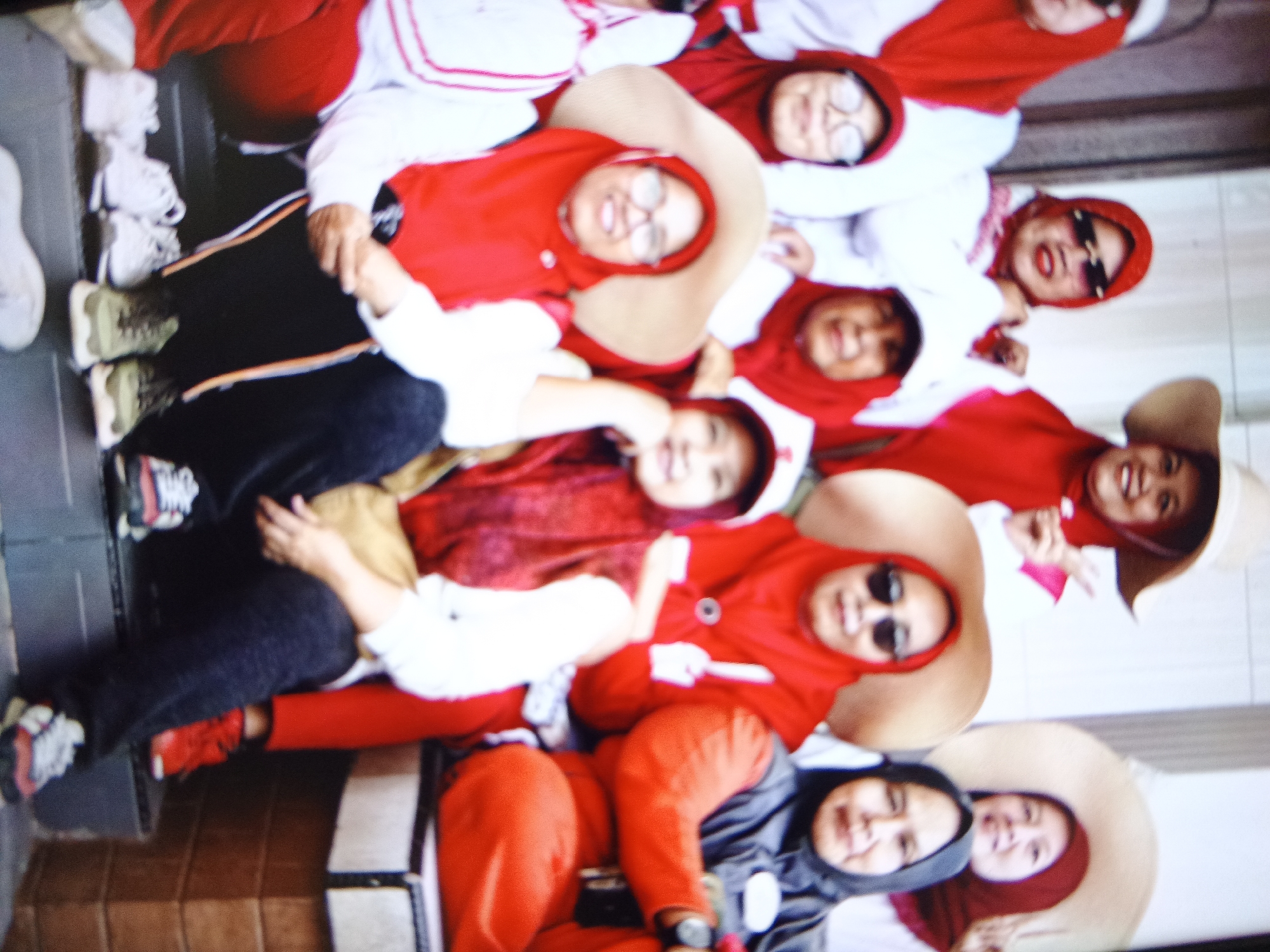 In a slightly blurry, angled outdoor shot taken in Warungkiara, Indonesia, a group of young people, likely students given their similar red and white attire, are lying down on a tiled surface. They appear to be posing for a photograph. The subjects are wearing red hooded jackets and some have large, light-colored hats. Their faces are mostly visible and some are smiling. Several individuals are wearing glasses or sunglasses. In the foreground, their legs and feet, clad in sneakers, are visible. The background shows a tiled wall.