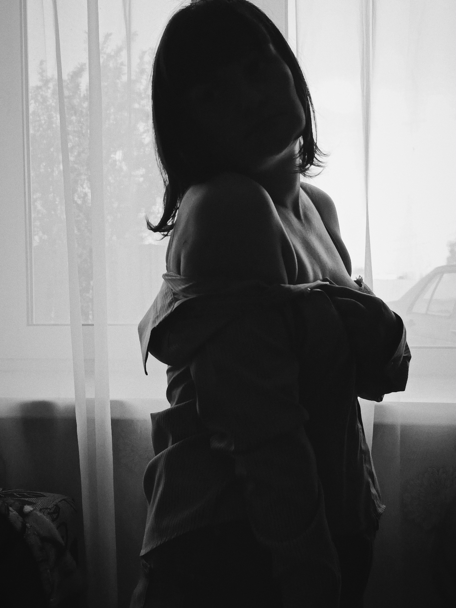 A black and white photograph shows a woman standing in front of a window. The woman is partially undressed, wearing a button-down shirt draped over her shoulders. Her face is obscured by shadow. The window is covered with a sheer curtain, and a tree and part of a car are visible outside. A chair or other upholstered furniture is partially visible in the lower left corner of the image. The location is listed as Unknown, Unknown.