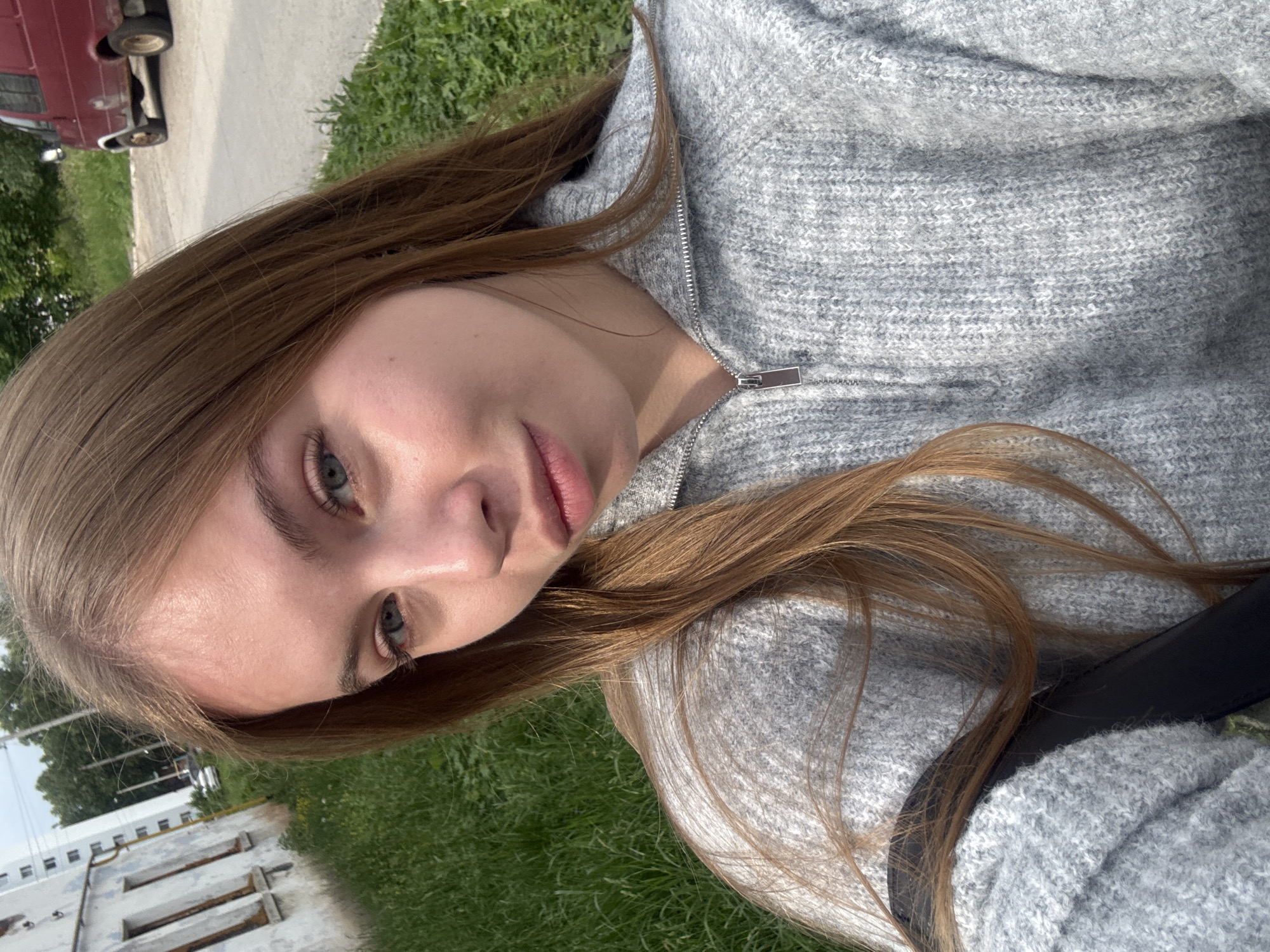 The image is a close-up, tilted selfie of a young woman with long, straight, light brown hair. She has fair skin, light-colored eyes (blue/grey), and a neutral expression as she looks directly at the camera. She is wearing a grey, knitted zippered hoodie or jacket, with a black strap, possibly from a bag, visible over her shoulder.
The setting is outdoors during the daytime, under what appears to be clear or partly cloudy skies, indicated by the bright, natural lighting. In the background, to the left of the woman, a partial view of a red vehicle (likely an older model car or van) is visible on an asphalt or dirt road. Green grass covers much of the ground in the mid-ground and foreground. In the distance, several white or light-colored multi-story buildings with windows are discernible, partially obscured by trees. The overall scene suggests a suburban or less urbanized environment, consistent with a town such as Chaikovsky, Russia. There is no visible text in the image.