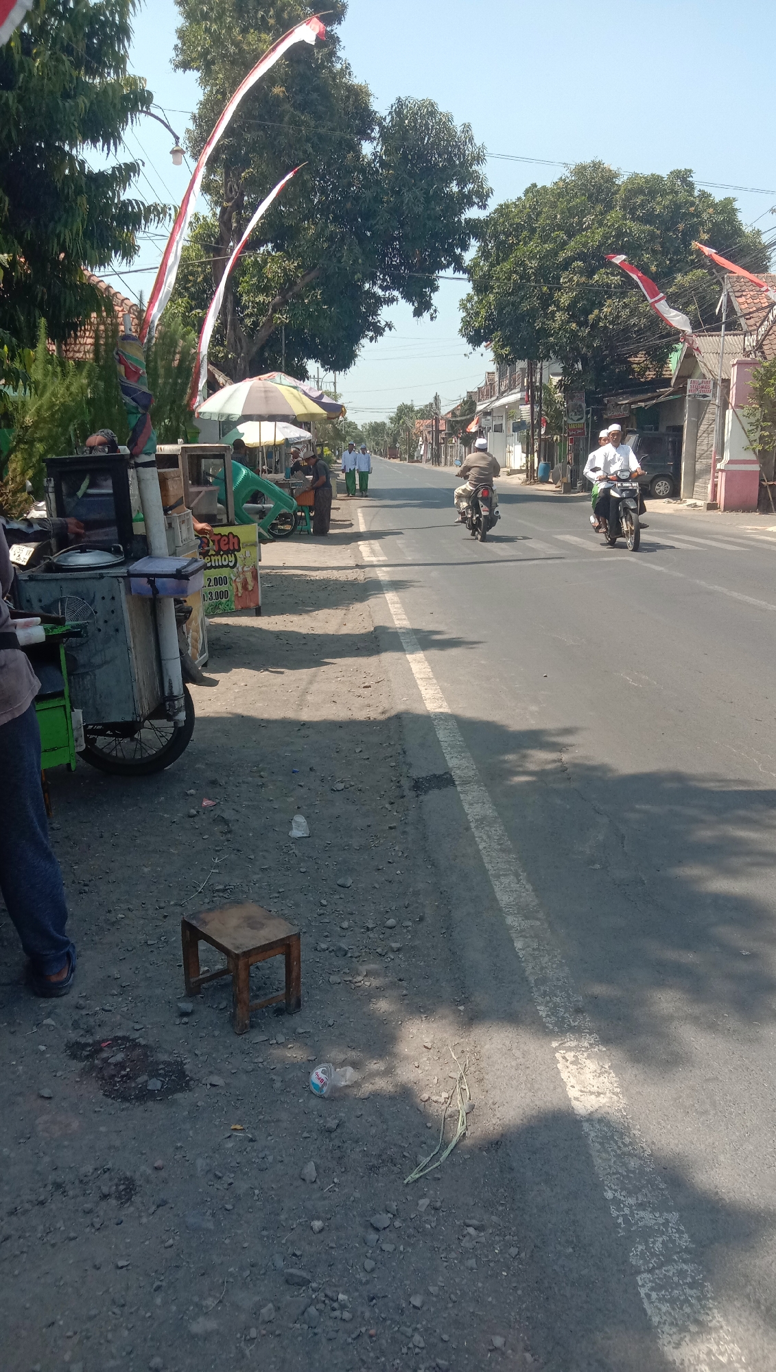 The image depicts a street scene in Karang Anyar, Indonesia. The foreground features a food cart with various items, including a green cooler, a metal serving tray, and a sign with prices. To the left of the cart, a person's legs and feet are visible, wearing dark pants and sandals. A small wooden stool sits on the gravelly ground, near a dark stain.
Further down the street, several people are visible. Two individuals in traditional attire are walking on the sidewalk. Two motorcycles are also present on the road. One motorcycle, ridden by a person wearing a white shirt and cap, is in the middle of the road. Another motorcycle, ridden by a person in a brown shirt and cap, is ahead of the first motorcycle and further to the right. Several trees line the street, some adorned with red and white flags, suggesting a festive occasion or national celebration. Buildings with visible roofs and some signage are present on the right side of the road. The sky is clear and blue, indicating a sunny day. The road surface is asphalt with white painted lines. The overall environment suggests a typical urban or semi-urban street in Indonesia.
