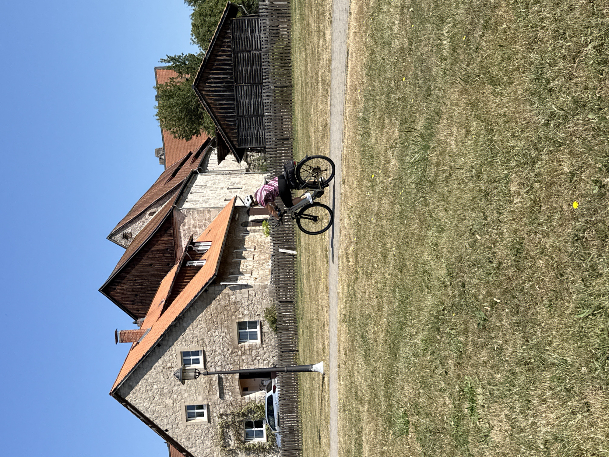 In Walkenried, Germany, a person wearing a pink and white striped shirt, black shorts, and a white helmet rides a bicycle on a paved path. The path runs through a dry, grassy field. Behind the field, a wooden fence separates it from a property with several buildings. The buildings appear to be old, constructed of stone and wood with red tile roofs. One building is a two-story stone structure with multiple windows and a prominent chimney. Adjacent to it is a large wooden barn-like structure. The sky is clear and blue. A light pole is visible near the stone building. A silver car is parked in front of the stone building.
