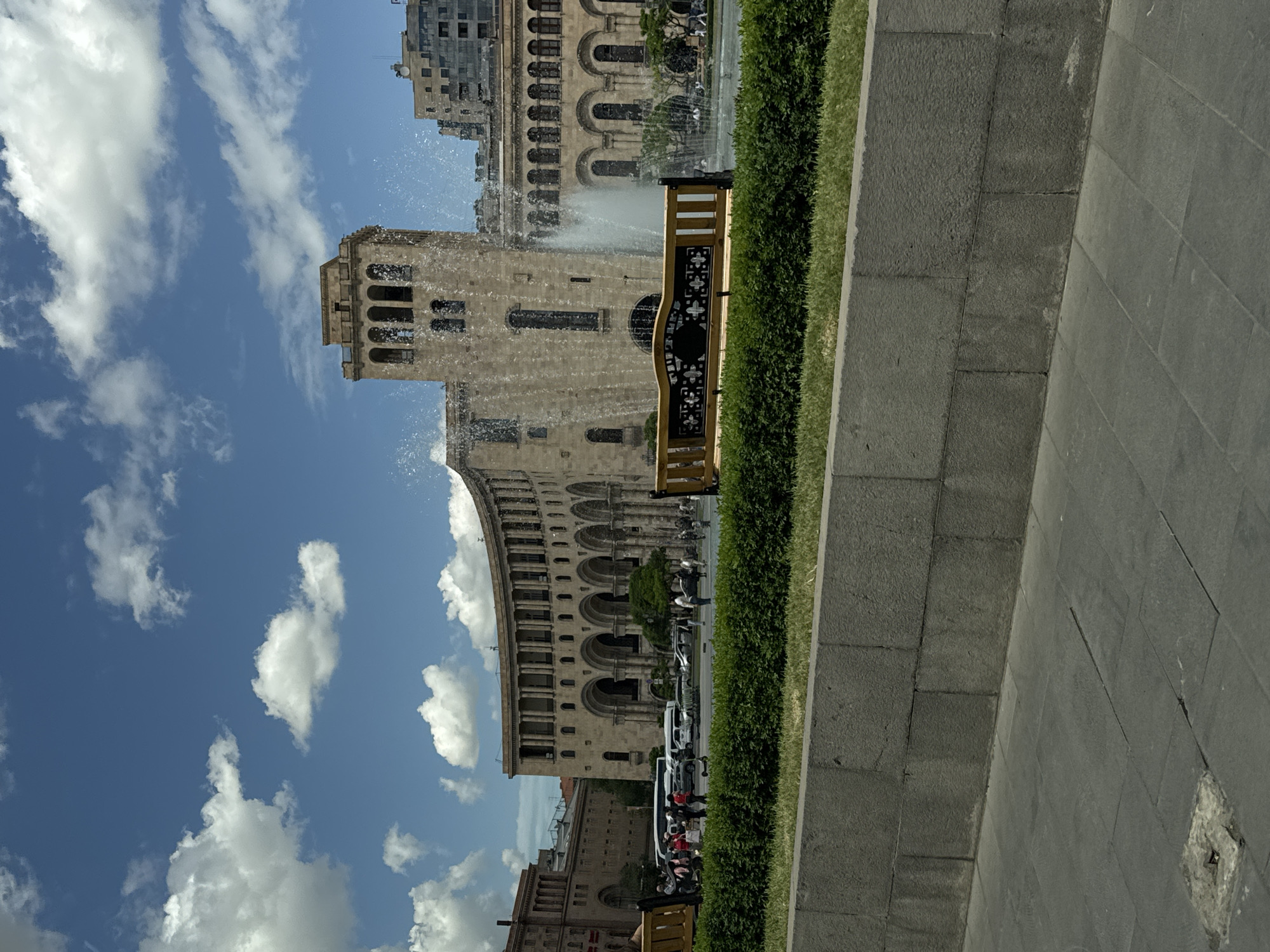 The image depicts a view of Republic Square in Yerevan, Armenia. The scene shows a grand stone building, identifiable as part of the National Gallery of Armenia, with its distinctive arched windows and a tower. A fountain is active in front of the building, with water jets arcing into the air. Pedestrians are visible on the sidewalk, and vehicles, possibly cars and buses, are parked or moving along the road in front of the building. A wooden bench is placed near a green hedge. The sky is bright blue with puffy white clouds. The foreground features a stone-tiled walkway.