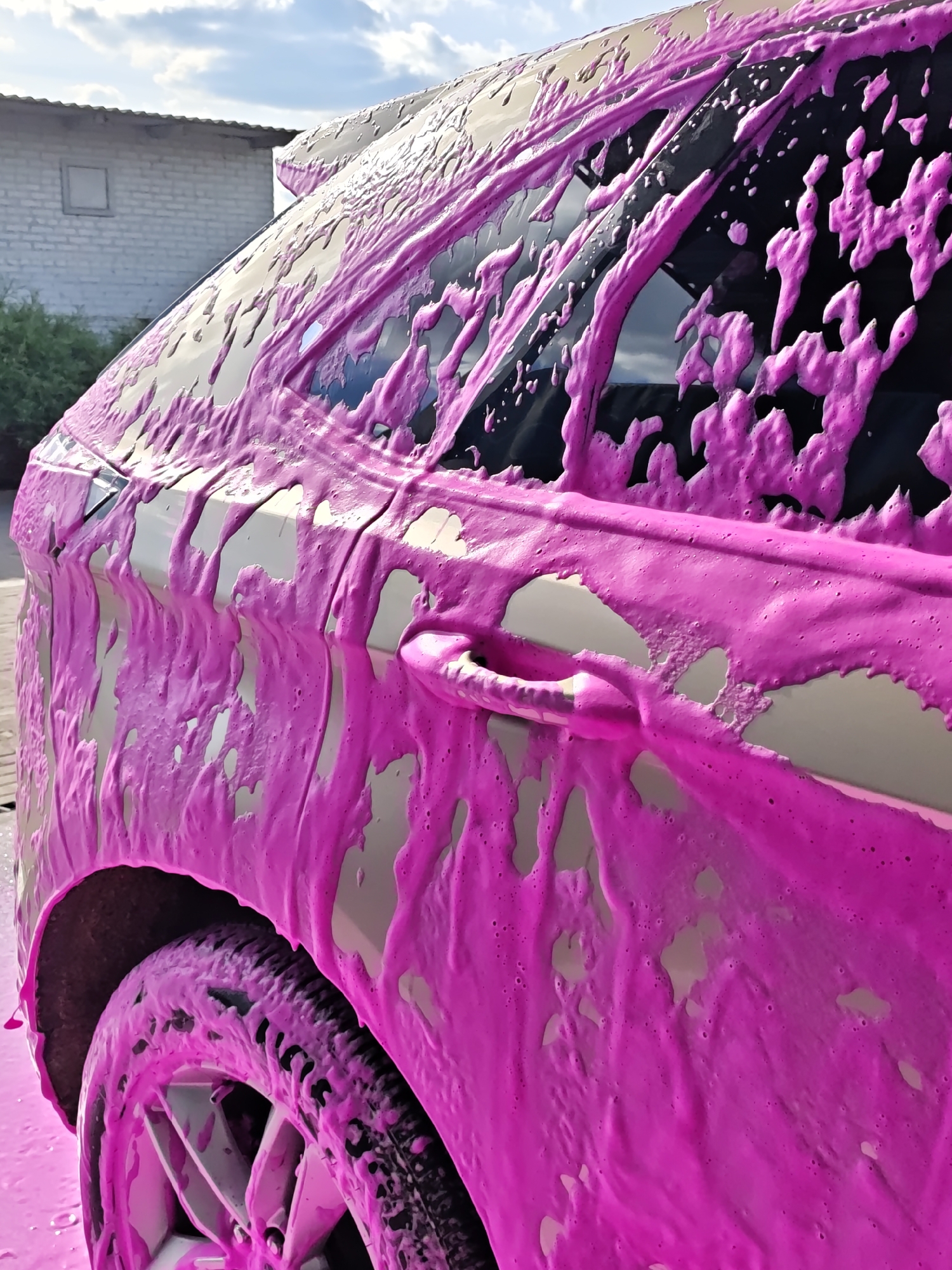 A close-up, brightly lit image shows the rear side of a car covered in pink soap suds. The car appears to be undergoing a wash. Details visible include the rear tire, door handle, and side window which are all heavily coated with the pink foam. In the background, a gray brick building with a small window is visible, along with some greenery. The location is Vinnytsia, Ukraine.