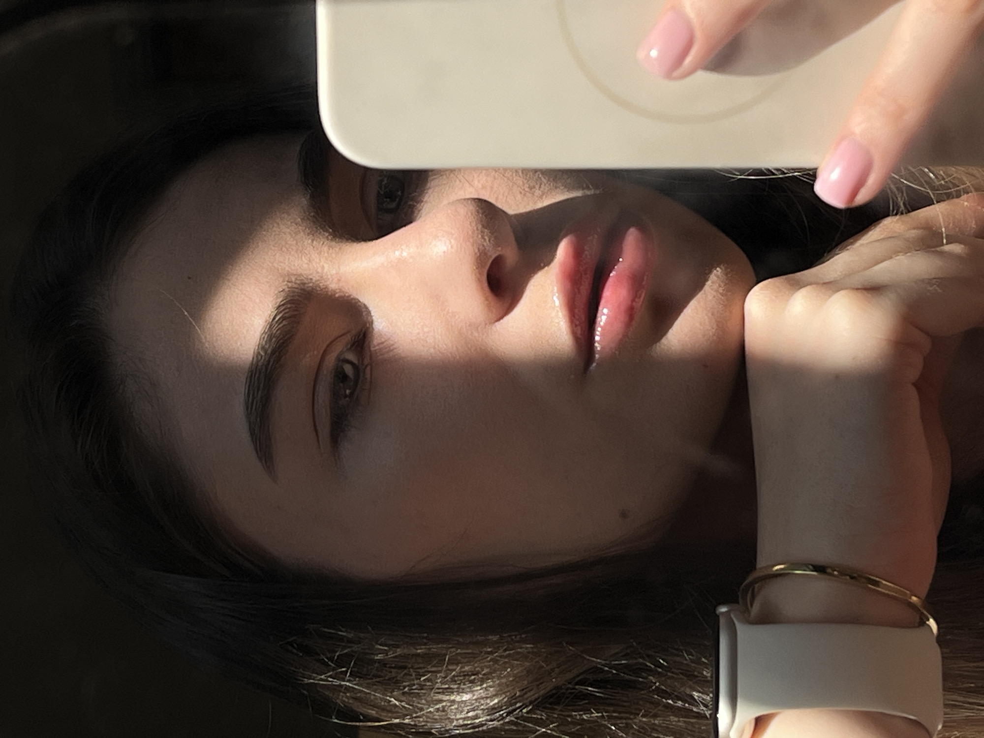 A person with brown hair is holding a smartphone and taking a selfie. The background is dark and undefined. The person is wearing a gold bracelet and a white smartwatch band. Their fingernails are painted a light pink. Light is illuminating the person's face. The location is Unknown, Unknown.