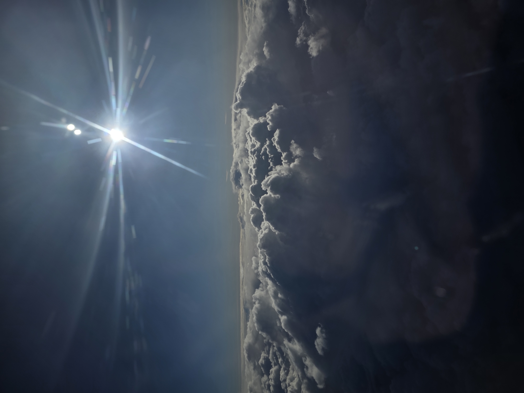 The image depicts a view from a high altitude, possibly from an airplane or a very high vantage point. On the left is a bright sun with a strong lens flare effect. The right side of the image shows a dense layer of clouds. The location in the city of Kommunarka, Russia is not discernible from the provided image.