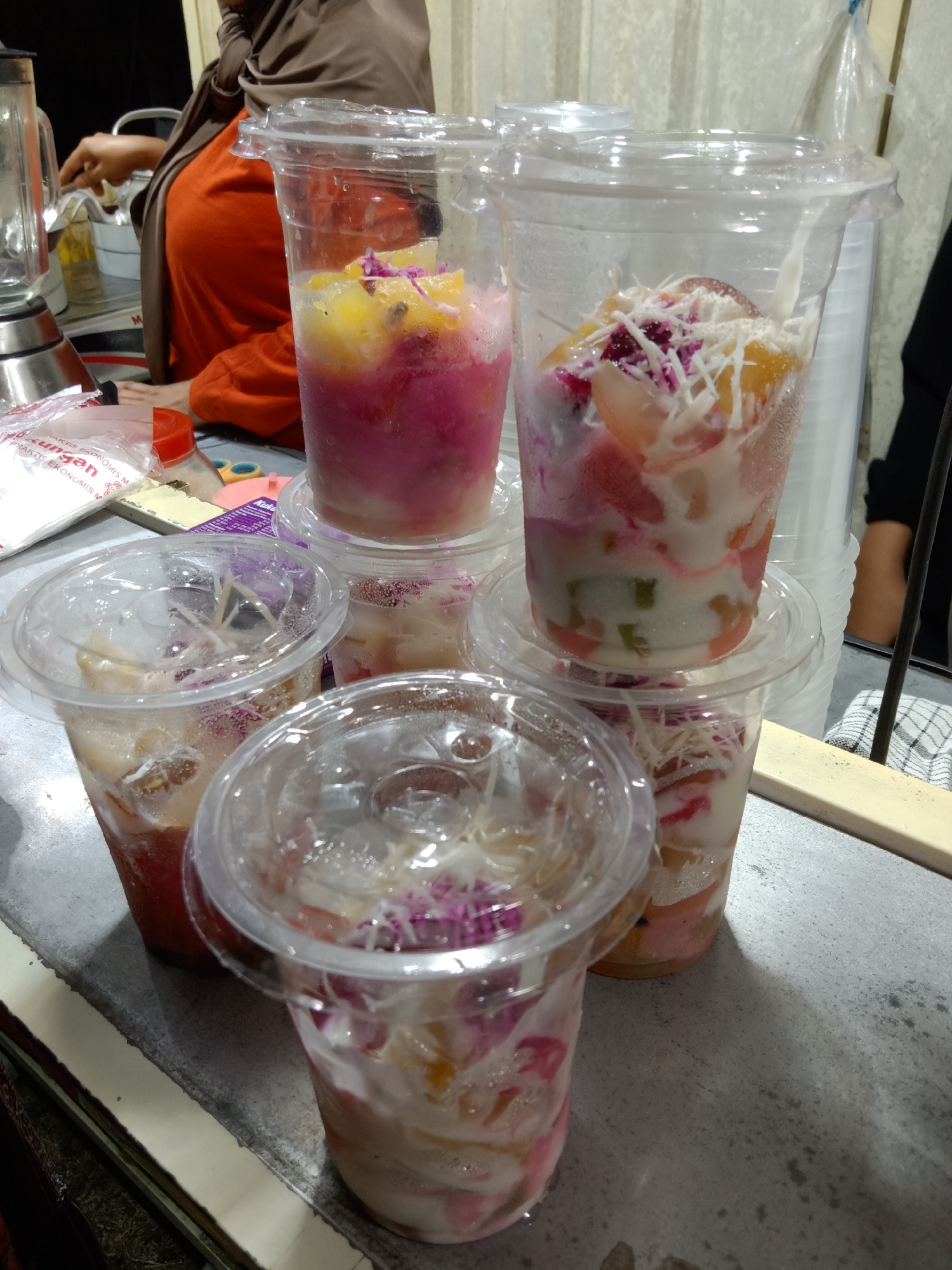 The image shows a close-up view of several plastic cups filled with a colorful fruit dessert, likely served at a street food stall. The cups are stacked and arranged on a grey countertop. Some cups contain chunks of pink and yellow fruit, layered with what appears to be shredded coconut or cheese, and possibly a creamy white liquid or yogurt. The lighting suggests an indoor setting, possibly a covered stall or a shop with overhead lighting. In the background, a person wearing an orange shirt and a brown hijab is partially visible, suggesting the vendor. The overall scene conveys a sense of a casual, perhaps bustling, food service environment in Stiaji, Indonesia.