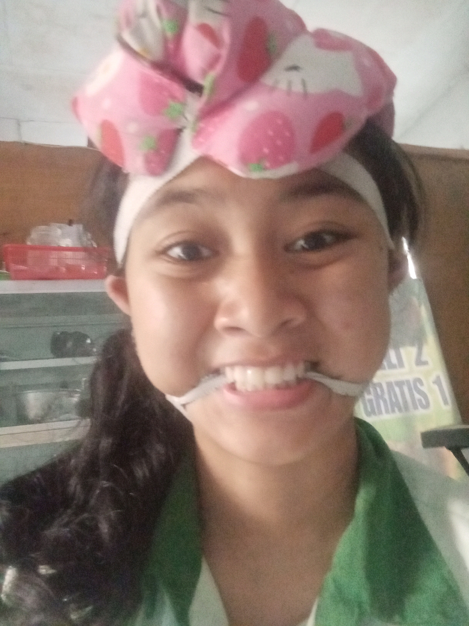 A young Indonesian woman, likely in Badas, Indonesia, is captured in a close-up selfie. She is wearing a pink headband adorned with a strawberry pattern. A white fabric strip is stretched across her mouth, holding her lips open to reveal her teeth. Her dark hair is visible on the left side of the frame. In the background, shelves are stocked with various items, including a red plastic container. A sign with text, partially obscured, is visible to the right. The lighting suggests an indoor environment.